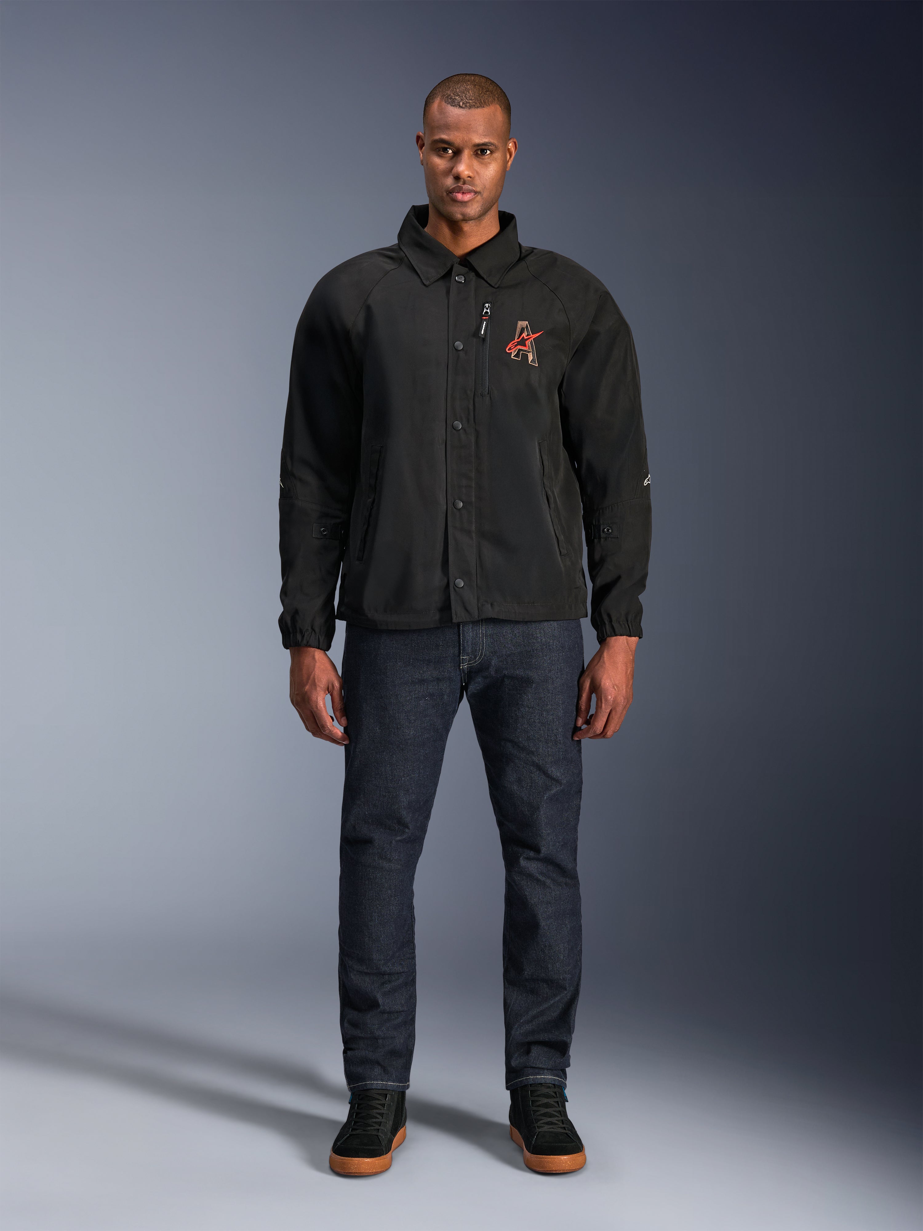 Chaqueta Alpinestars Revved WR en color negro, vestida por un modelo con vaqueros azul oscuro y zapatillas negras, vista frontal, con cierre de botones a presión y logotipo bordado en 3D en el pecho, equipamiento de motociclismo urbano contemporáneo.