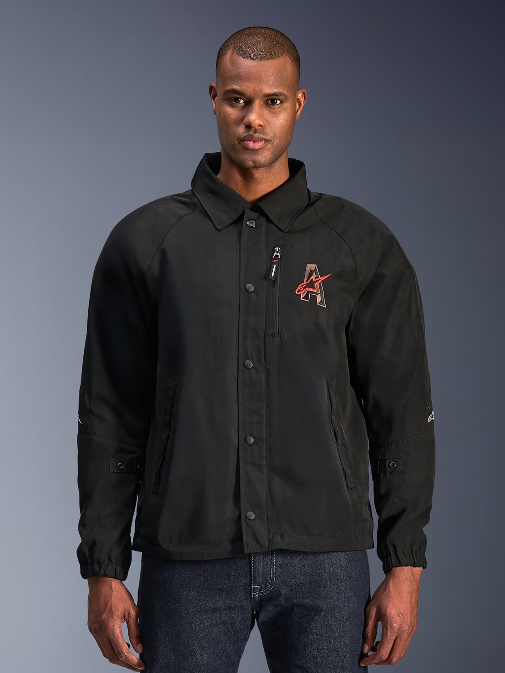 Alpinestars Revved WR Jacket, chaqueta de moto negra con cuello tipo camisa y cierre frontal de botones a presión, logotipo 'A' rojo bordado en el pecho, bolsillo vertical con cremallera en el pecho, puños elásticos, diseño de chaqueta tipo coaches urbana