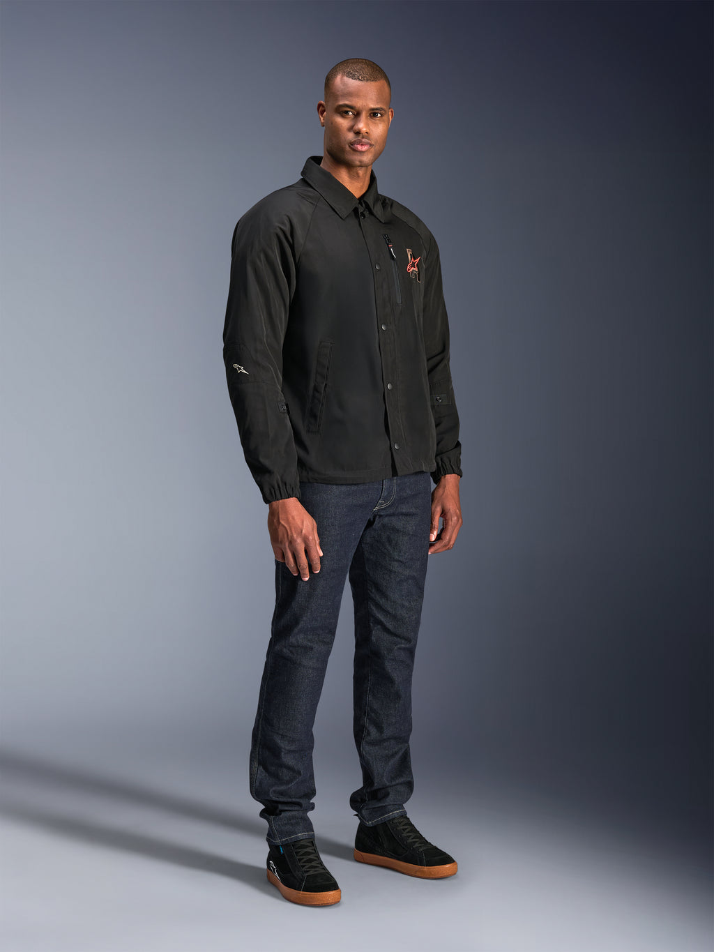 Chaqueta Alpinestars Revved WR en color negro, chaqueta de moto urbana de estilo entrenador usada por un modelo con vaqueros azul oscuro y zapatillas negras, con cierre frontal de botones a presión y logotipo bordado, diseño minimalista y deportivo para la conducción en carretera.