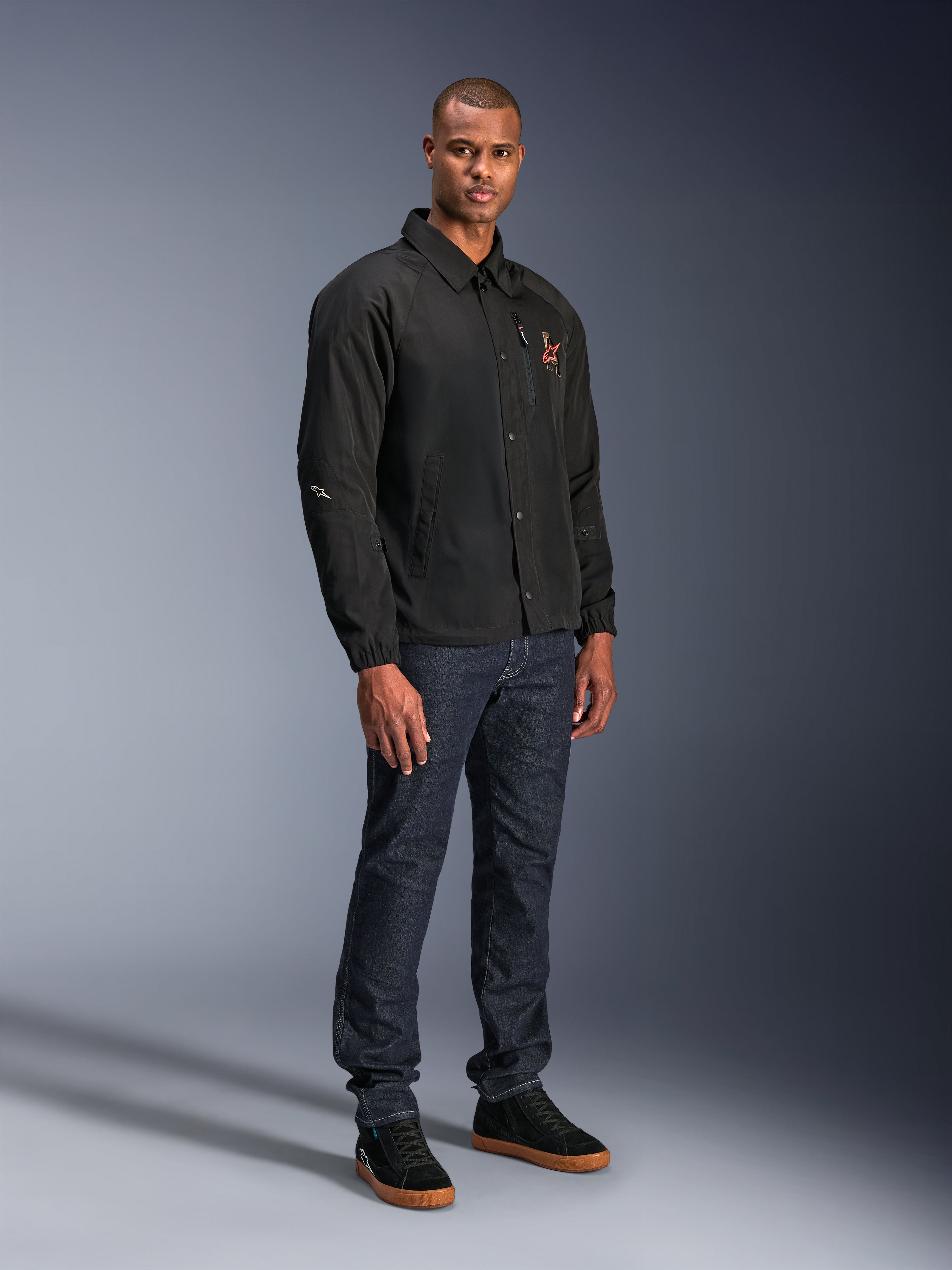 Chaqueta Alpinestars Revved WR en color negro, chaqueta de moto urbana de estilo entrenador usada por un modelo con vaqueros azul oscuro y zapatillas negras, con cierre frontal de botones a presión y logotipo bordado, diseño minimalista y deportivo para la conducción en carretera.