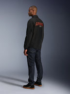Chaqueta Alpinestars Revved WR, negra, con un gran logotipo de Alpinestars bordado en 3D en color naranja en la espalda, vista trasera de tres cuartos de un modelo con vaqueros azul oscuro y zapatillas negras, chaqueta de moto urbana contemporánea con silueta de chaqueta de entrenador