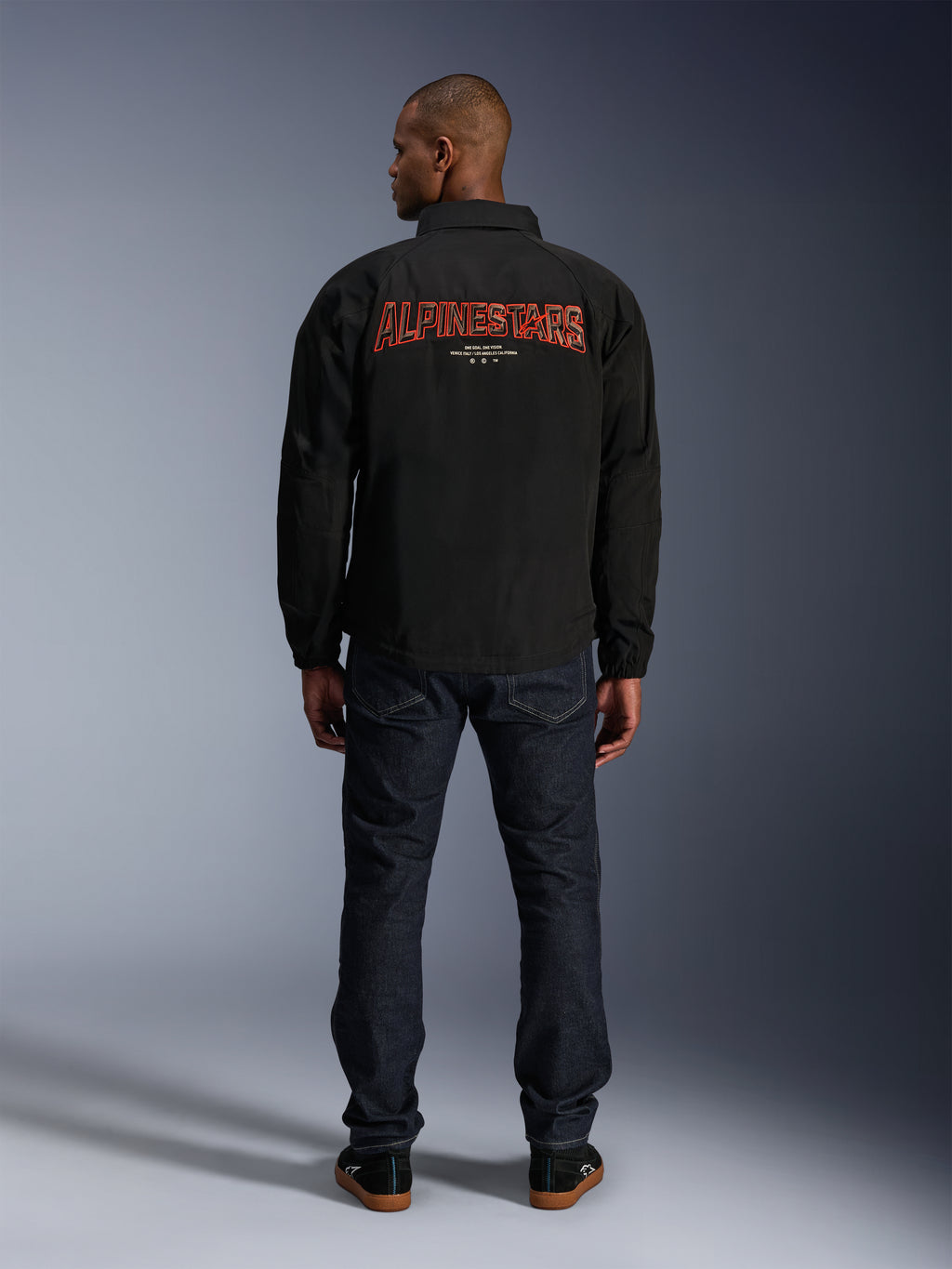 Chaqueta Alpinestars Revved WR, negra, vista trasera con un gran bordado rojo de 'ALPINESTARS' con efecto 3D, chaqueta urbana estilo entrenador, combinada con vaqueros de denim oscuro y zapatillas negras, ropa de motociclismo minimalista
