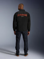 Chaqueta Alpinestars Revved WR, negra, vista trasera con un gran bordado rojo de 'ALPINESTARS' con efecto 3D, chaqueta urbana estilo entrenador, combinada con vaqueros de denim oscuro y zapatillas negras, ropa de motociclismo minimalista