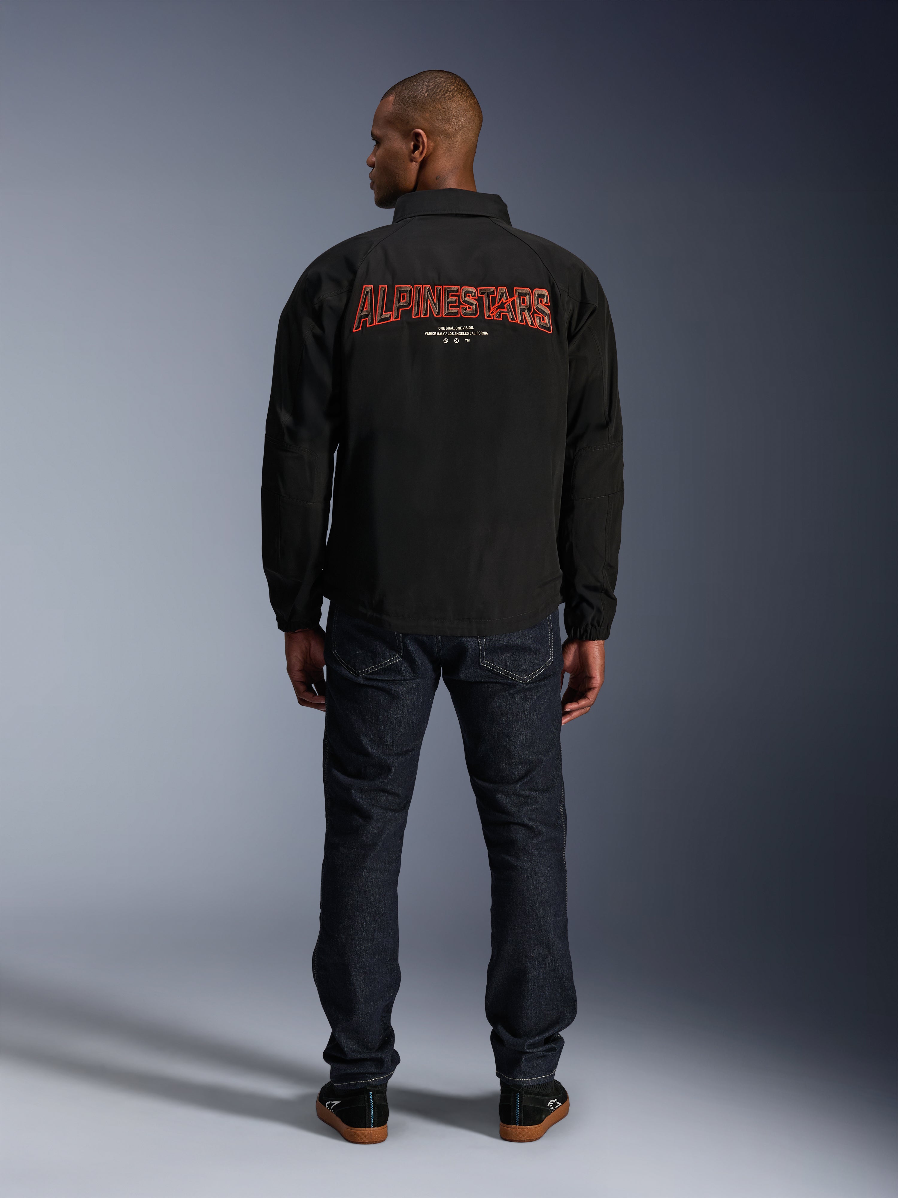 Chaqueta Alpinestars Revved WR, negra, vista trasera con un gran bordado rojo de 'ALPINESTARS' con efecto 3D, chaqueta urbana estilo entrenador, combinada con vaqueros de denim oscuro y zapatillas negras, ropa de motociclismo minimalista