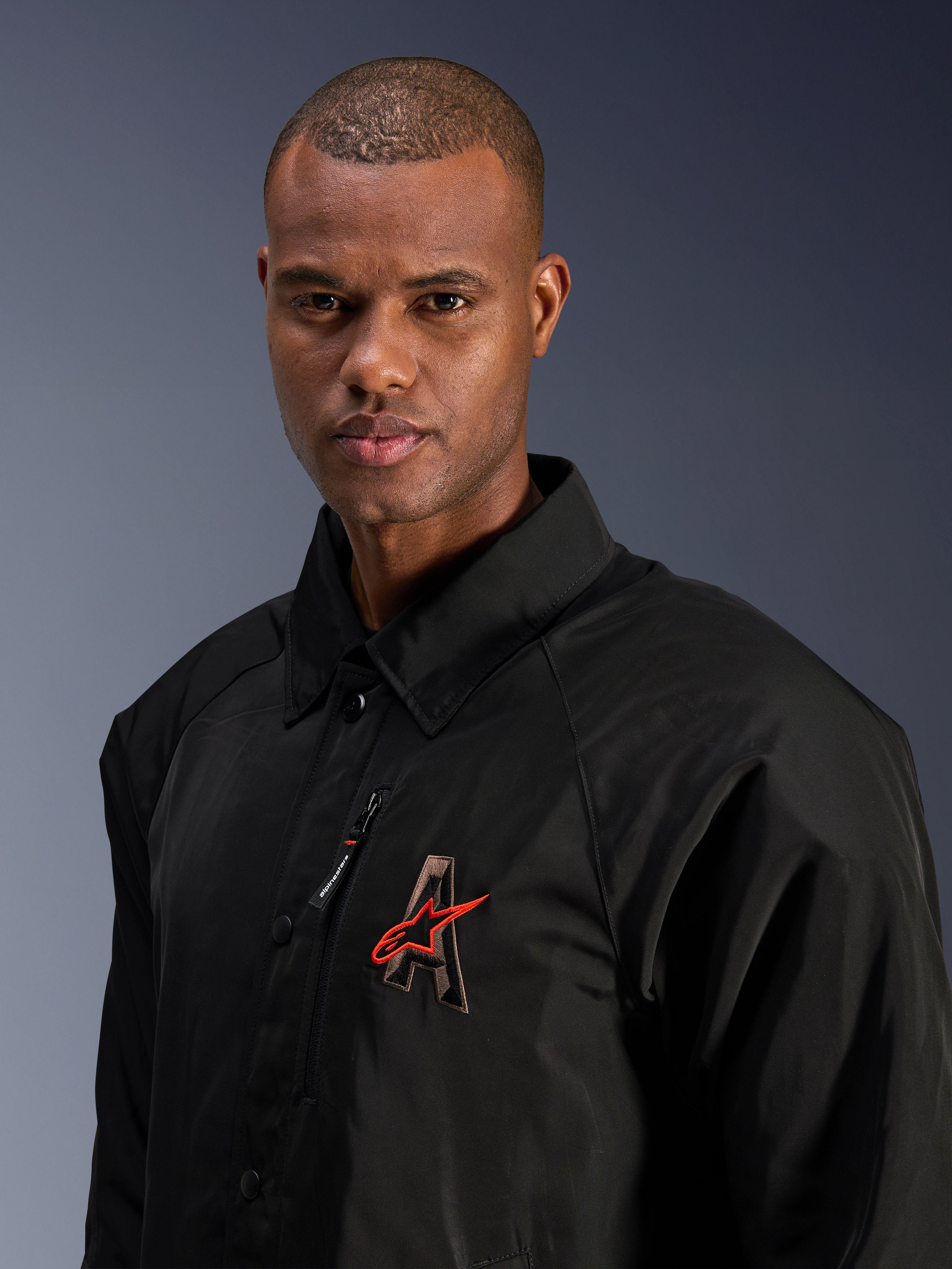Alpinestars Revved WR Jacket, negra, primer plano de un hombre con la chaqueta de motociclista de satén resistente al agua, con el logotipo 'A' bordado en 3D y la estrella roja de Alpinestars, bolsillo con cremallera en el pecho y diseño con cuello para conducción urbana contemporánea.