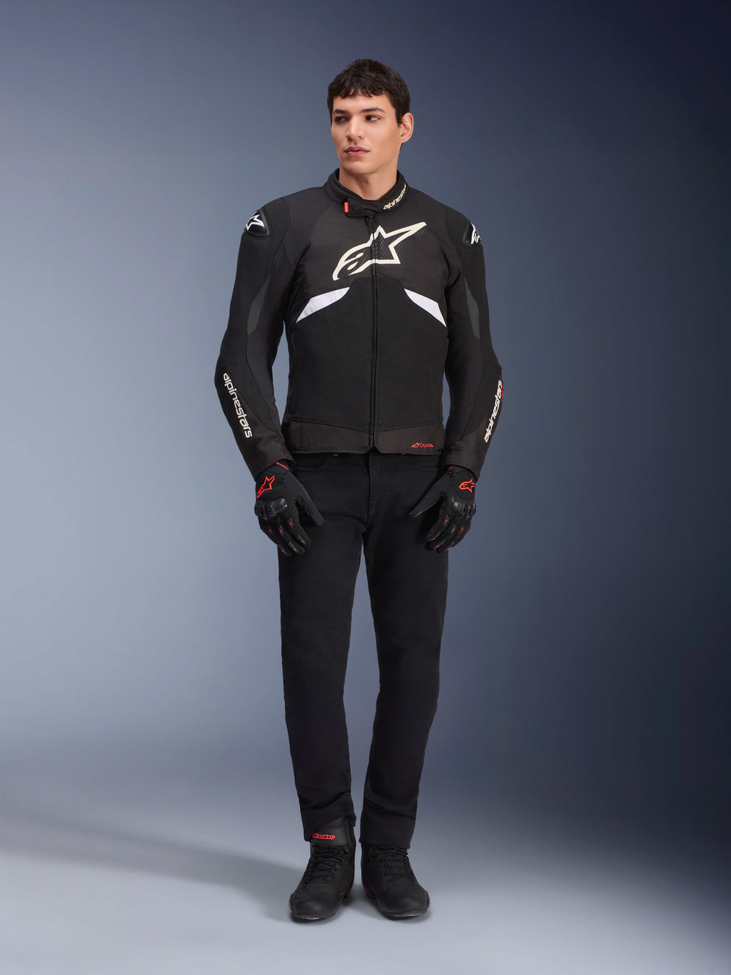 Modelo masculino luciendo la chaqueta Alpinestars T-GP R V3 Drystar&reg; en negro y blanco, una chaqueta deportiva textil e impermeable con un gran logotipo blanco de Alpinestars en el pecho, combinada con pantalones y guantes negros; retrato de estudio profesional que resalta su estilizada silueta deportiva.