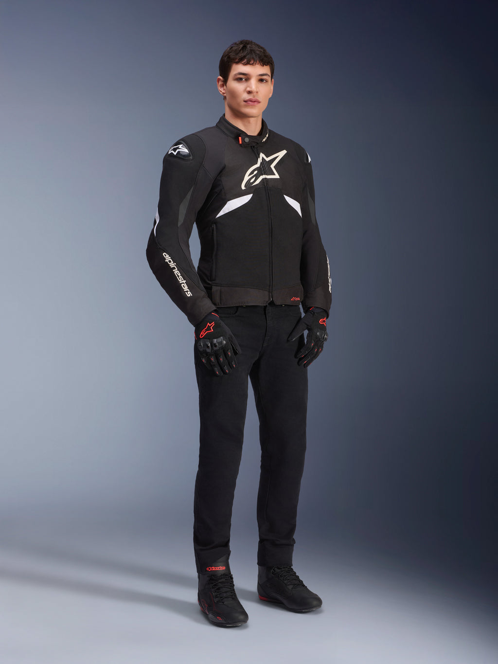 Chaqueta Alpinestars T-GP R V3 Drystar® en negro y blanco, vestida por un modelo con guantes y pantalones de moto negros, con un logotipo blanco en el pecho, detalles blancos en las mangas y deslizadores protectores en los hombros, chaqueta deportiva textil impermeable para conducción en carretera