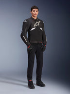 Chaqueta Alpinestars T-GP R V3 Drystar® en negro y blanco, vestida por un modelo con guantes y pantalones de moto negros, con un logotipo blanco en el pecho, detalles blancos en las mangas y deslizadores protectores en los hombros, chaqueta deportiva textil impermeable para conducción en carretera