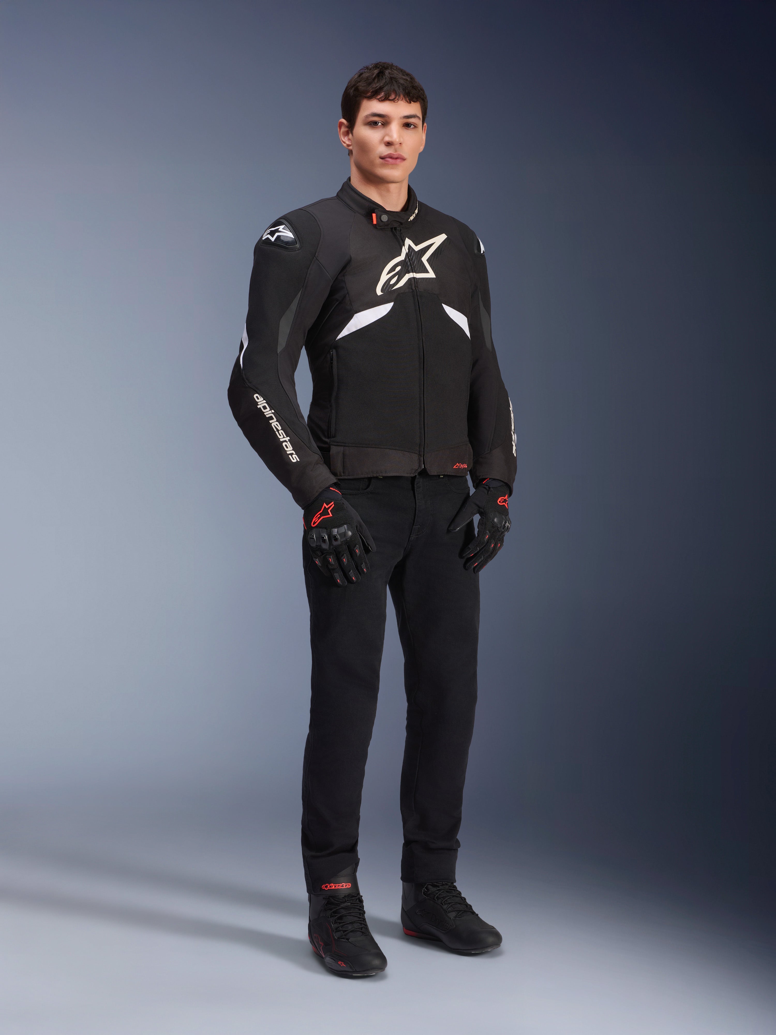 Chaqueta Alpinestars T-GP R V3 Drystar® en negro y blanco, vestida por un modelo con guantes y pantalones de moto negros, con un logotipo blanco en el pecho, detalles blancos en las mangas y deslizadores protectores en los hombros, chaqueta deportiva textil impermeable para conducción en carretera