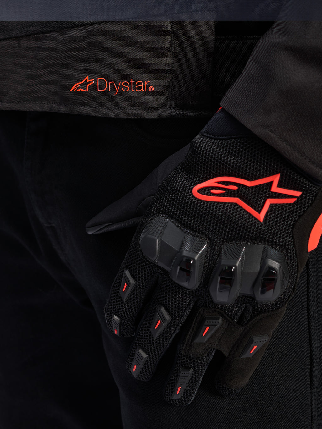 Primer plano de la cintura de la chaqueta Alpinestars T-GP R V3 Drystar® con el logotipo Drystar en rojo, mostrada con un guante de moto negro con el logotipo de la estrella de Alpinestars en rojo, tejido de malla y protección de nudillos reforzada.