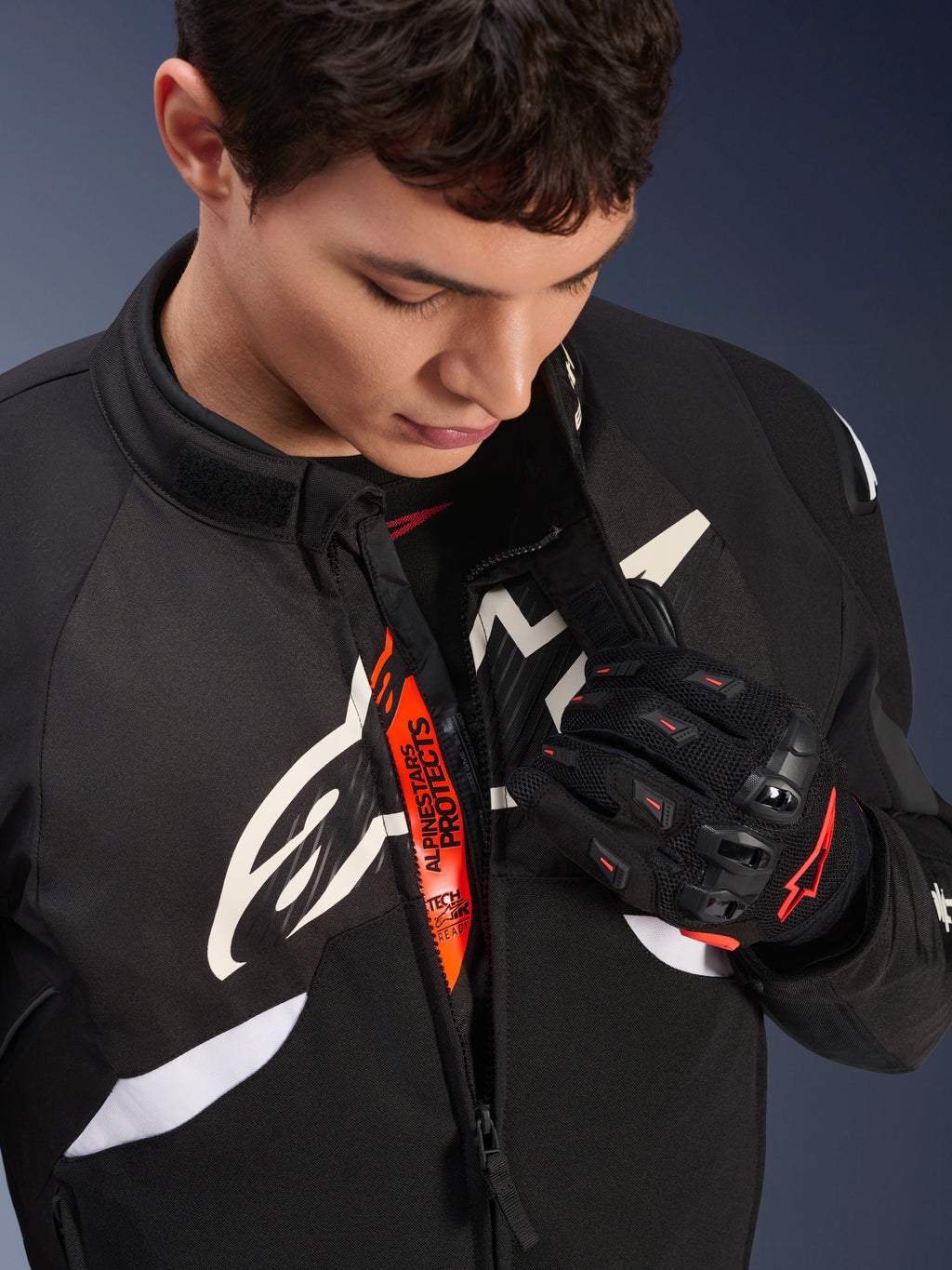 Chaqueta Alpinestars T-GP R V3 Drystar®, chaqueta textil deportiva para moto en blanco y negro, parcialmente abierta revelando la cinta interna naranja 'Alpinestars Protects', usada con guantes de malla negros y rojos, equipamiento de protección derivado de la competición.
