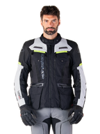 Hombre con la chaqueta Alpinestars Bogotá Pro Drystar®, en colores negro, gris hielo y amarillo flúor, una chaqueta de moto touring de aventura para todas las estaciones, con diseño impermeable, múltiples bolsillos y logotipo vertical, combinada con guantes negros y pantalones de motorista grises