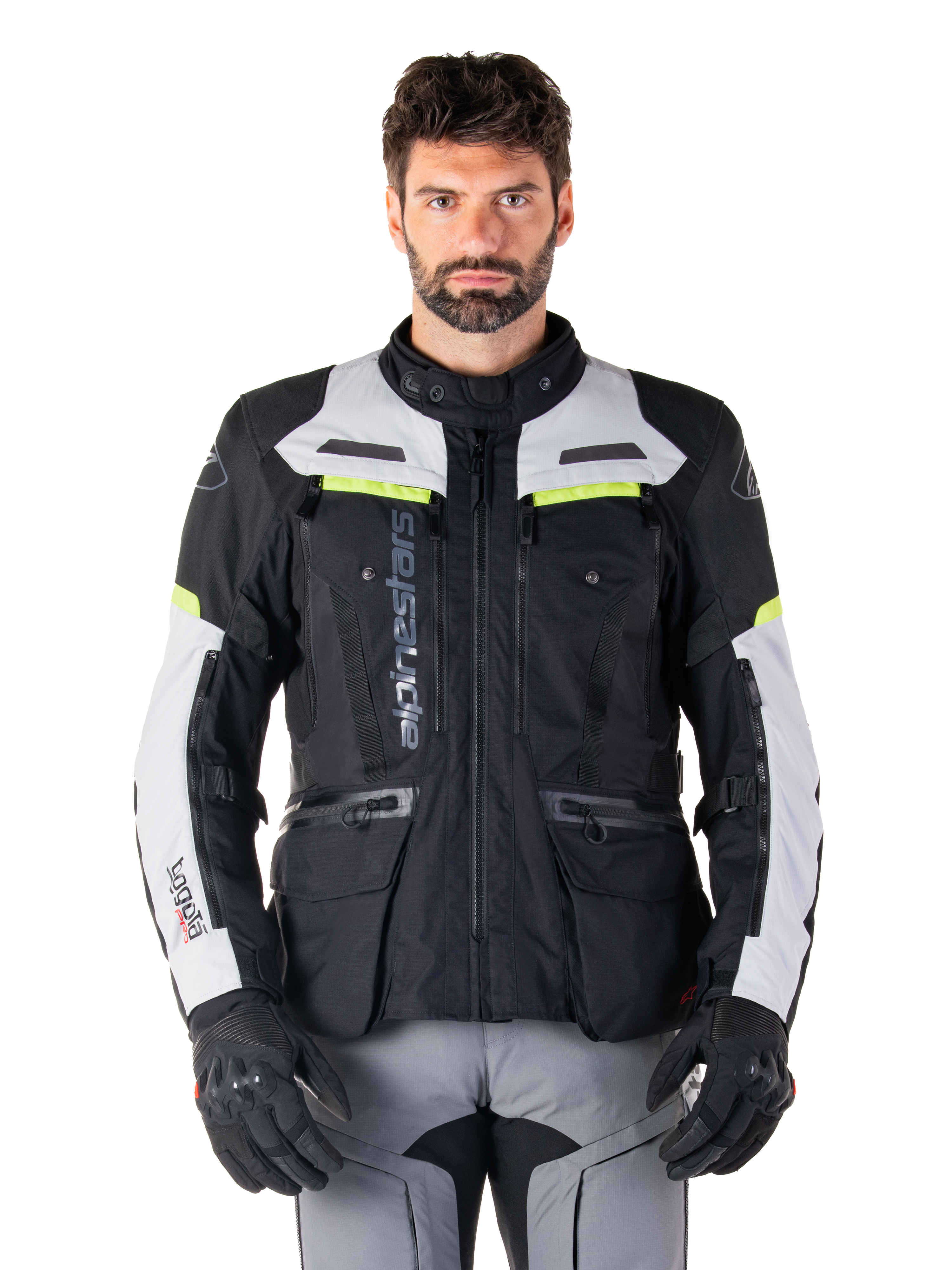 Hombre con la chaqueta Alpinestars Bogotá Pro Drystar®, en colores negro, gris hielo y amarillo flúor, una chaqueta de moto touring de aventura para todas las estaciones, con diseño impermeable, múltiples bolsillos y logotipo vertical, combinada con guantes negros y pantalones de motorista grises
