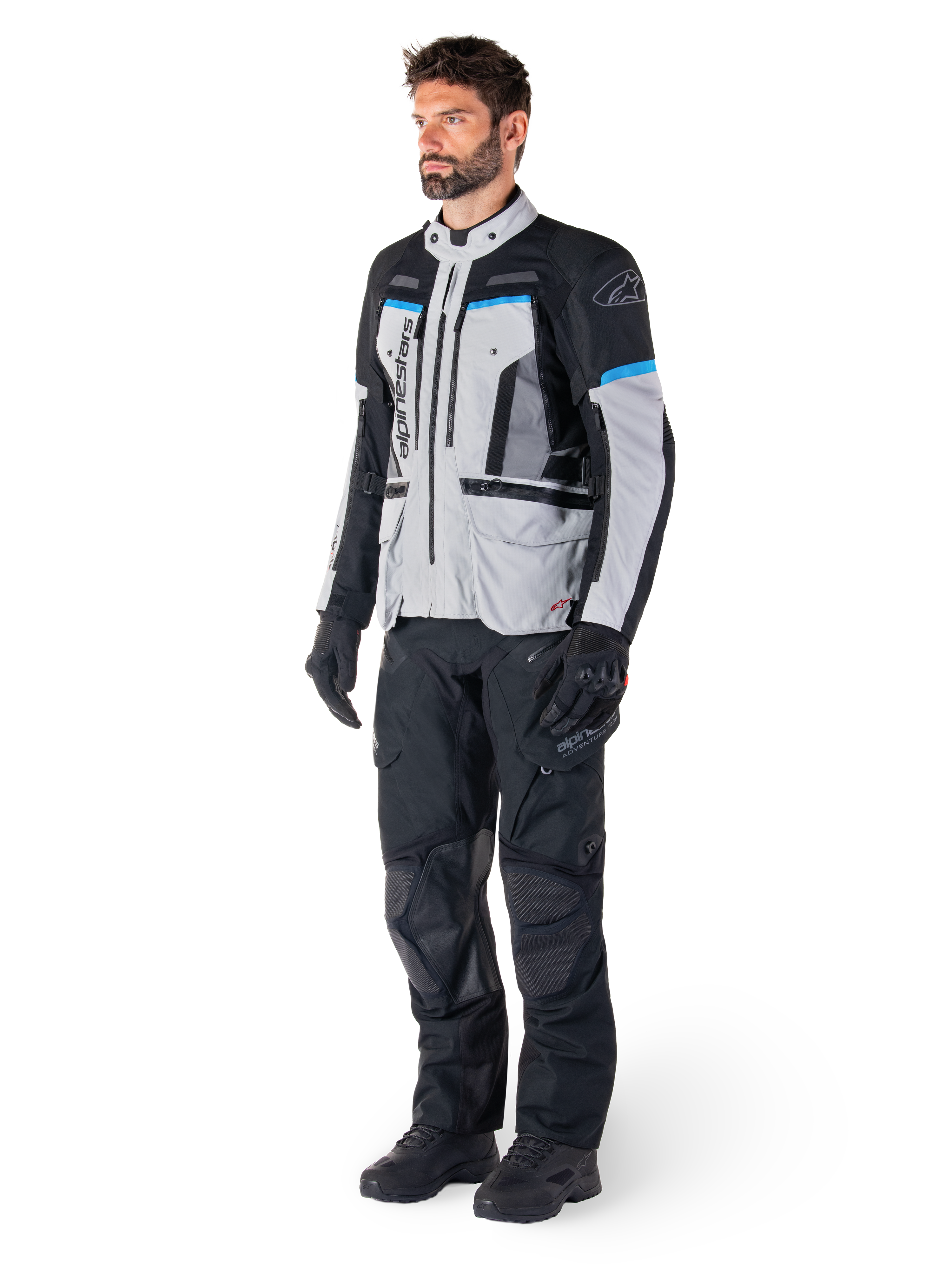 Male rider wearing Alpinestars Bogotá Pro Drystar® Jacket in Ice Gray, Black, and Cyan, paired with black technical riding pants and gloves, adventure touring gear for all-weather performance.