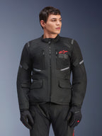 Chaqueta Alpinestars Andes Air Drystar®, chaqueta de moto touring negra, logo de estrella roja en el pecho, con paneles de ventilación de malla y detalles reflectantes, usada por un modelo masculino con guantes de moto negros, equipamiento de aventura para dos estaciones