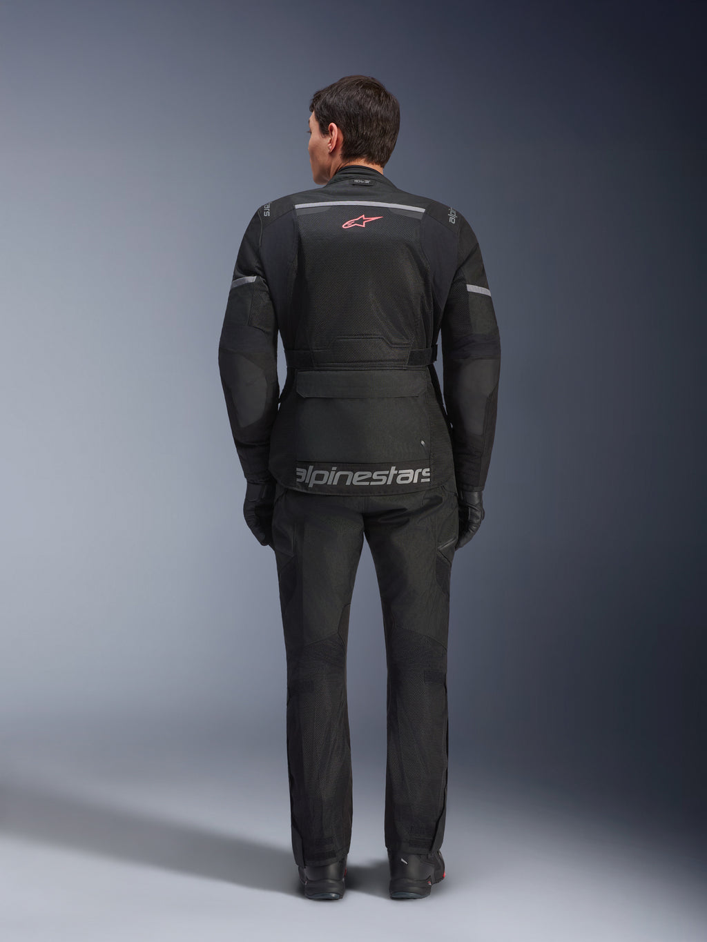 Vista trasera de un motorista con la chaqueta Alpinestars Andes Air Drystar® en color negro, con amplios paneles de ventilación de malla en la espalda, logotipo de Alpinestars en rojo y detalles reflectantes, combinada con pantalones y guantes de moto a juego para adventure touring.