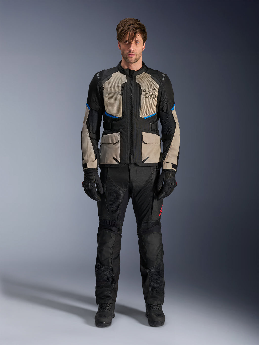 Modelo con chaqueta Alpinestars Andes Air Drystar®, chaqueta de moto touring en color negro, gris laurel y azul brillante, combinada con pantalones y guantes negros a juego, con amplios paneles de ventilación de malla en el pecho y las mangas para touring de aventura en climas cálidos.