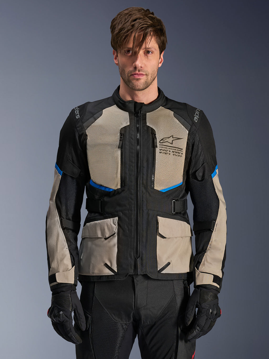 Chaqueta Alpinestars Andes Air Drystar, chaqueta de turismo de aventura, negro, gris laurel y azul brillante, usada por un modelo con guantes negros, con amplios paneles de malla en el pecho para flujo de aire, bolsillos de carga frontales y detalles en azul, equipo de protección para mototurismo