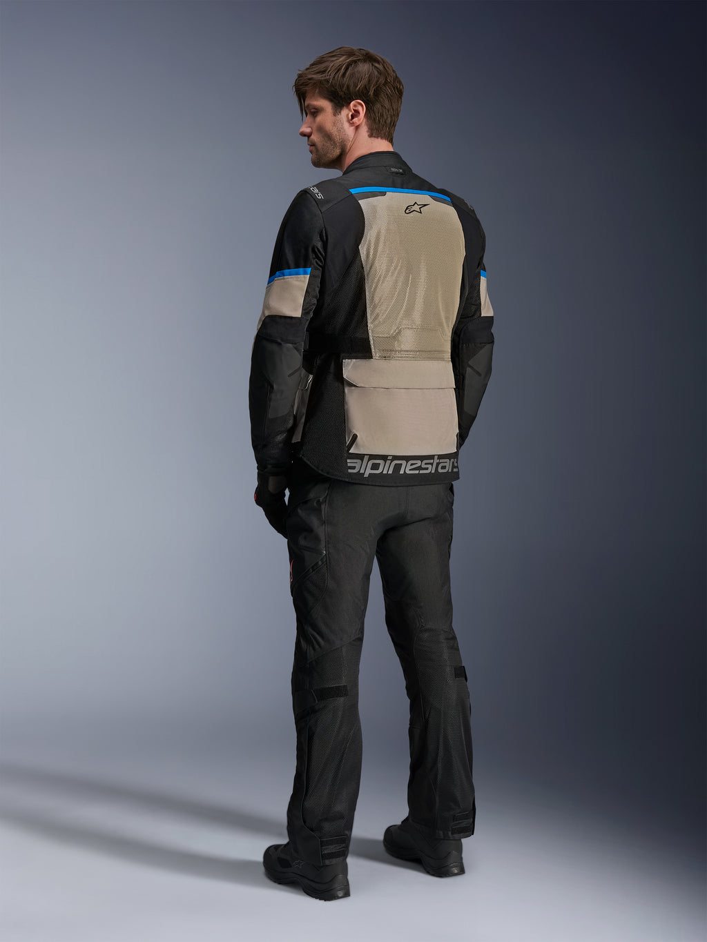 Chaqueta Alpinestars Andes Air Drystar®, negro, gris laurel y azul brillante, vista trasera que muestra un amplio panel de ventilación de malla en la espalda, logotipo en el dobladillo inferior, combinada con pantalones y botas de moto negros.