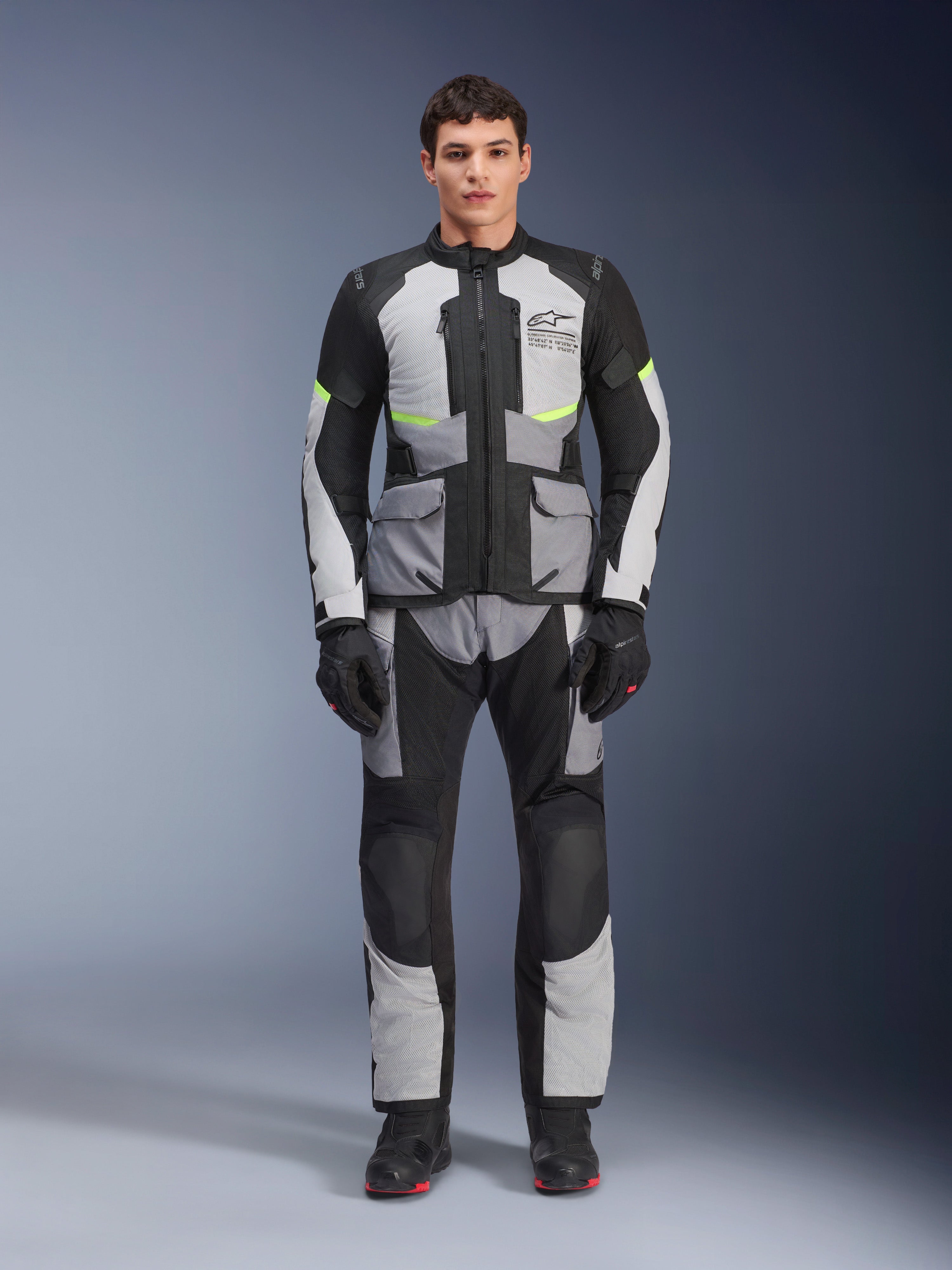 Chaqueta Alpinestars Andes Air Drystar® en gris hielo, gris oscuro y negro, usada por un modelo masculino con pantalones de motociclismo y guantes a juego, diseño de turismo de aventura con grandes bolsillos frontales y detalles en amarillo fluorescente, vista frontal de estudio