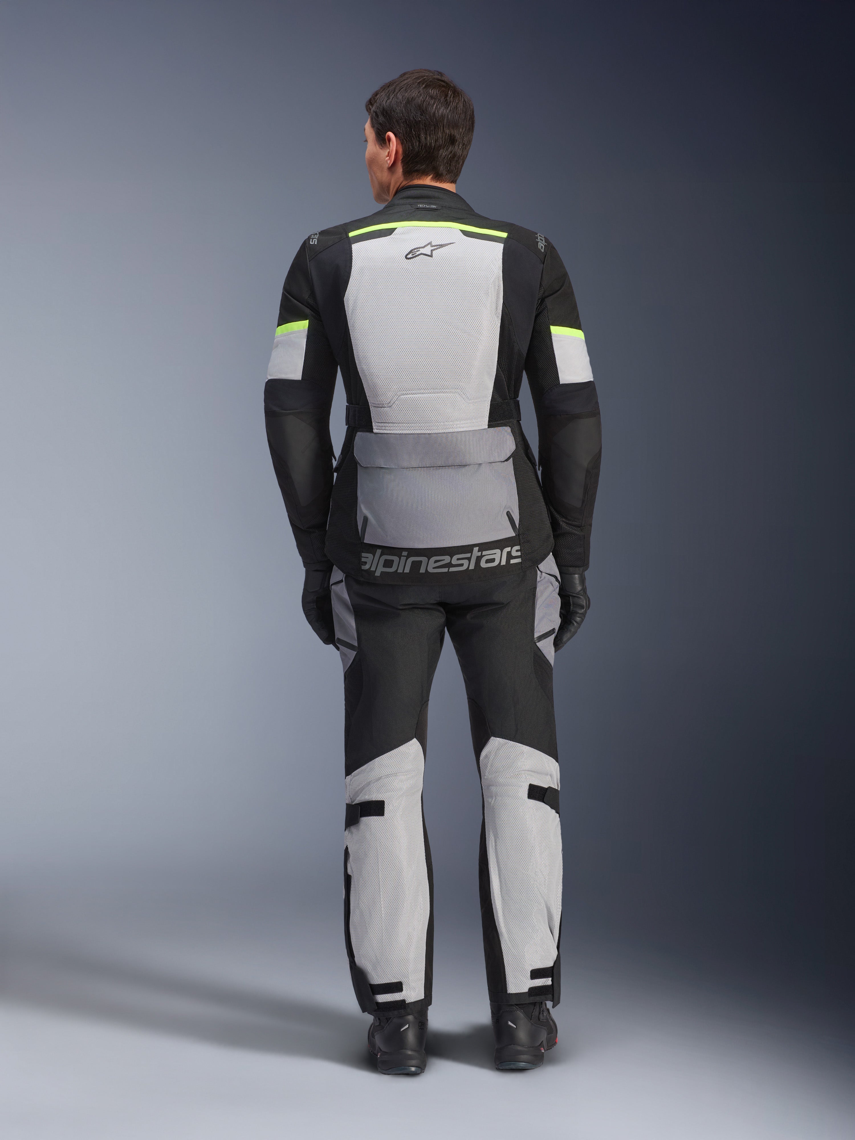 Chaqueta y pantalones Alpinestars Andes Air Drystar®, conjunto de touring de aventura en Ice Gray Dark Gray Black, vista trasera del piloto con la equipación completa y detalles en amarillo neón, con amplios paneles de ventilación de malla y logotipo de Alpinestars.