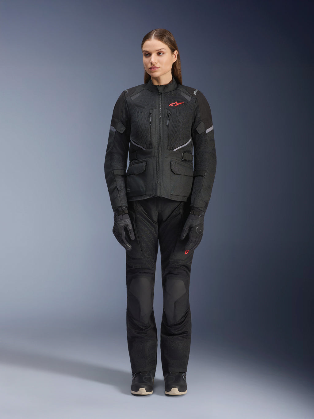 Modelo femenina con la chaqueta Alpinestars Stella Andes Air Drystar® en color negro, combinada con pantalones de turismo y guantes negros a juego, con amplios paneles de ventilación de malla, detalles de logotipos en rojo y elementos reflectantes, fotografía de estudio profesional de equipamiento de motociclismo para mujer.