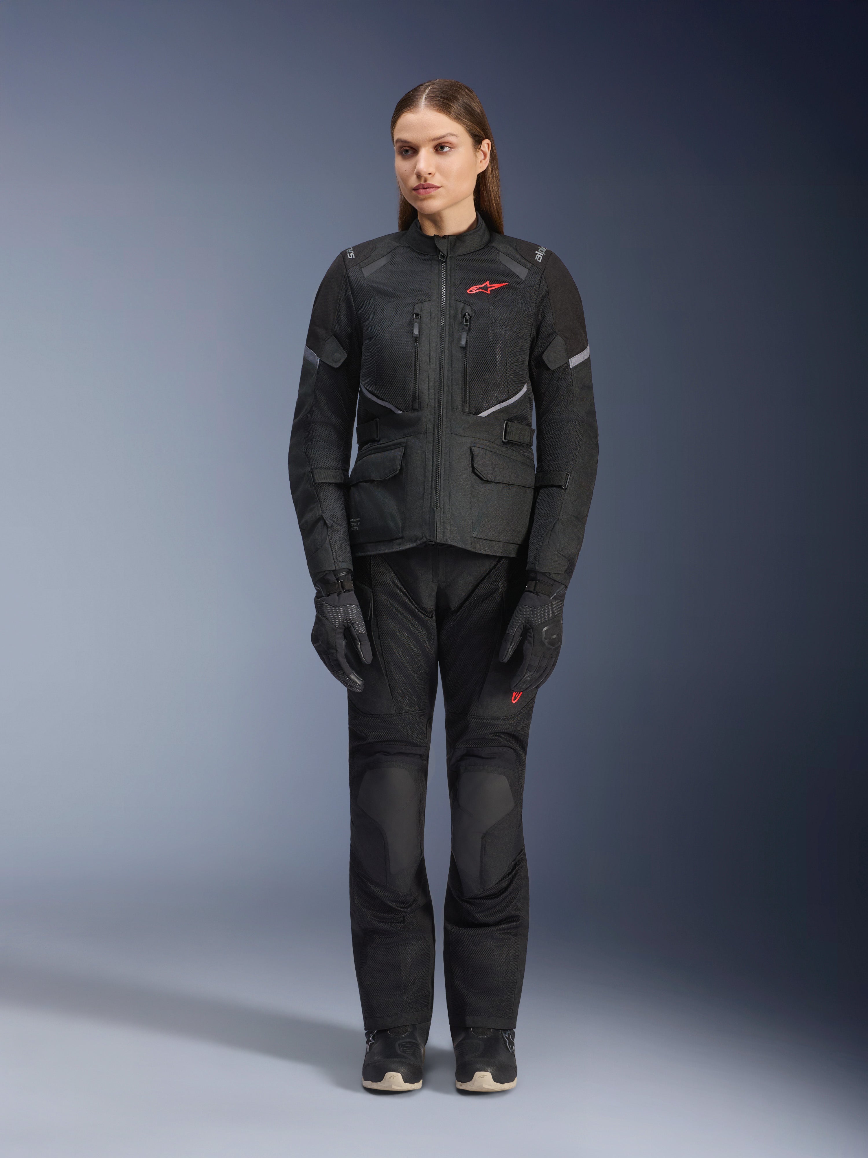 Modelo femenina con la chaqueta Alpinestars Stella Andes Air Drystar® en color negro, combinada con pantalones de turismo y guantes negros a juego, con amplios paneles de ventilación de malla, detalles de logotipos en rojo y elementos reflectantes, fotografía de estudio profesional de equipamiento de motociclismo para mujer.
