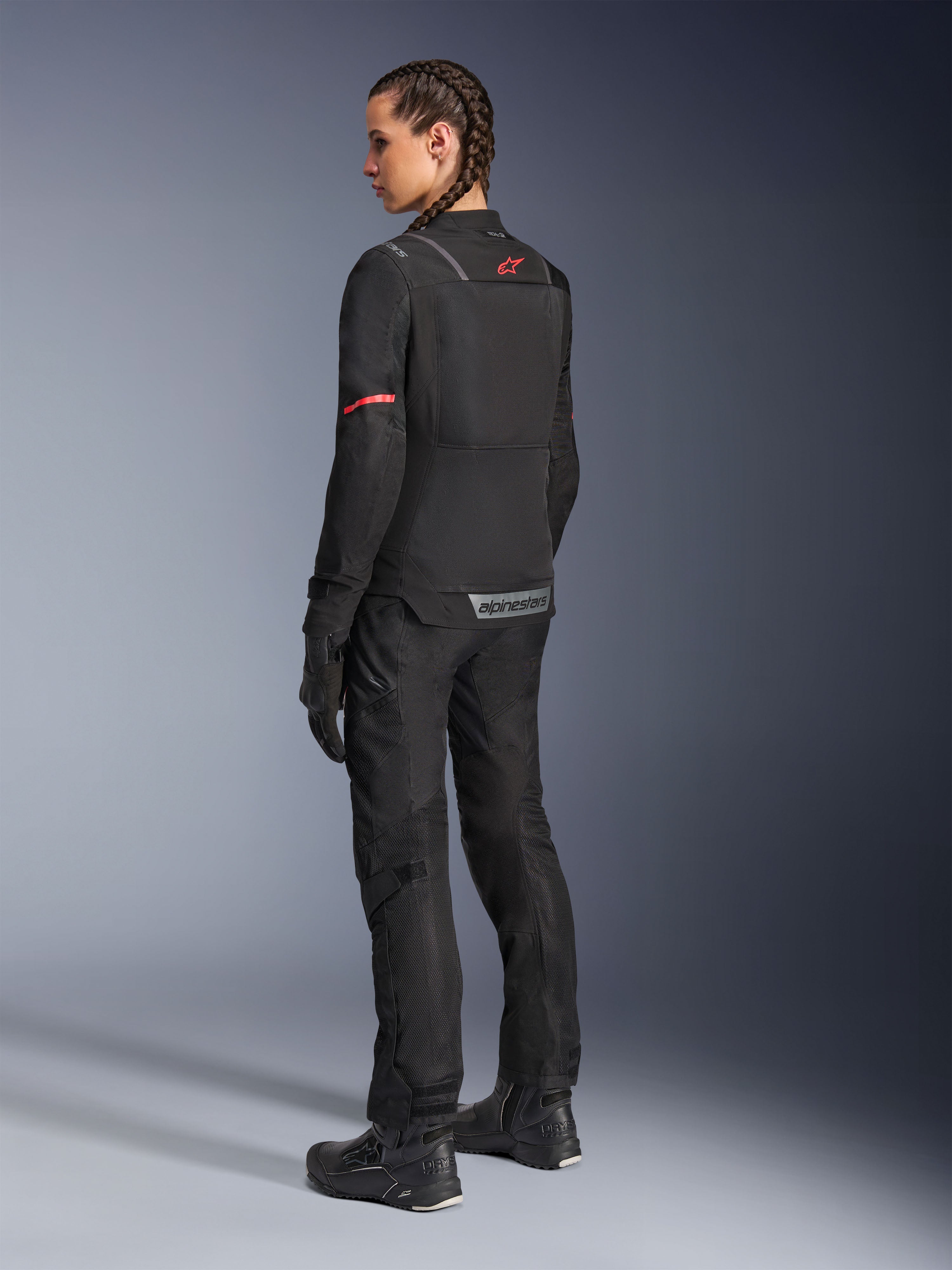 Chaqueta Alpinestars Stella ST-2 Air, chaqueta de moto para mujer, negra con detalles rojos en las mangas y la parte superior de la espalda, vista posterior en modelo que viste pantalones y botas de moto negros a juego, con marca Alpinestars reflectante y construcción textil moderna.