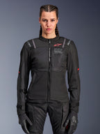 Alpinestars Stella ST-2 Air Jacket, chaqueta de moto para mujer, negra, vista frontal en modelo, con paneles de malla, logotipo rojo en el pecho y marca Alpinestars Exploration Equipment, usada con guantes negros y pantalones de moto