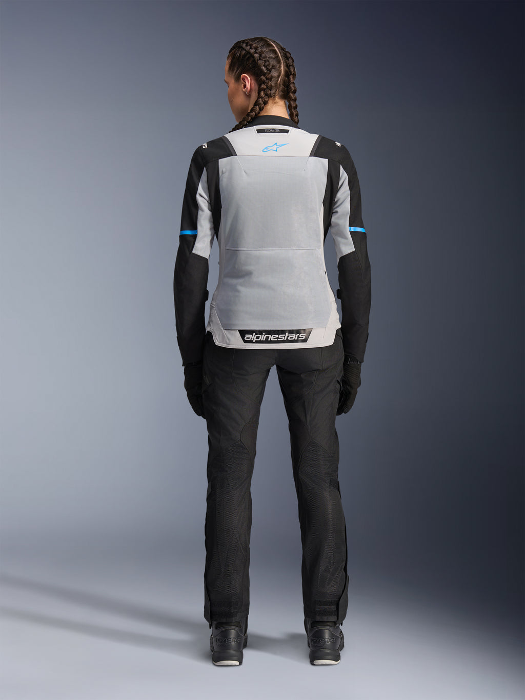 Chaqueta Alpinestars Stella ST-2 Air, Ice Gray Black, vista trasera en modelo femenina, con un gran panel de malla gris claro en la espalda, mangas negras con detalles de franjas azules, logotipo de Alpinestars en la parte inferior de la espalda, combinada con pantalones técnicos de conducción y botas negras, equipamiento de touring de verano para moto