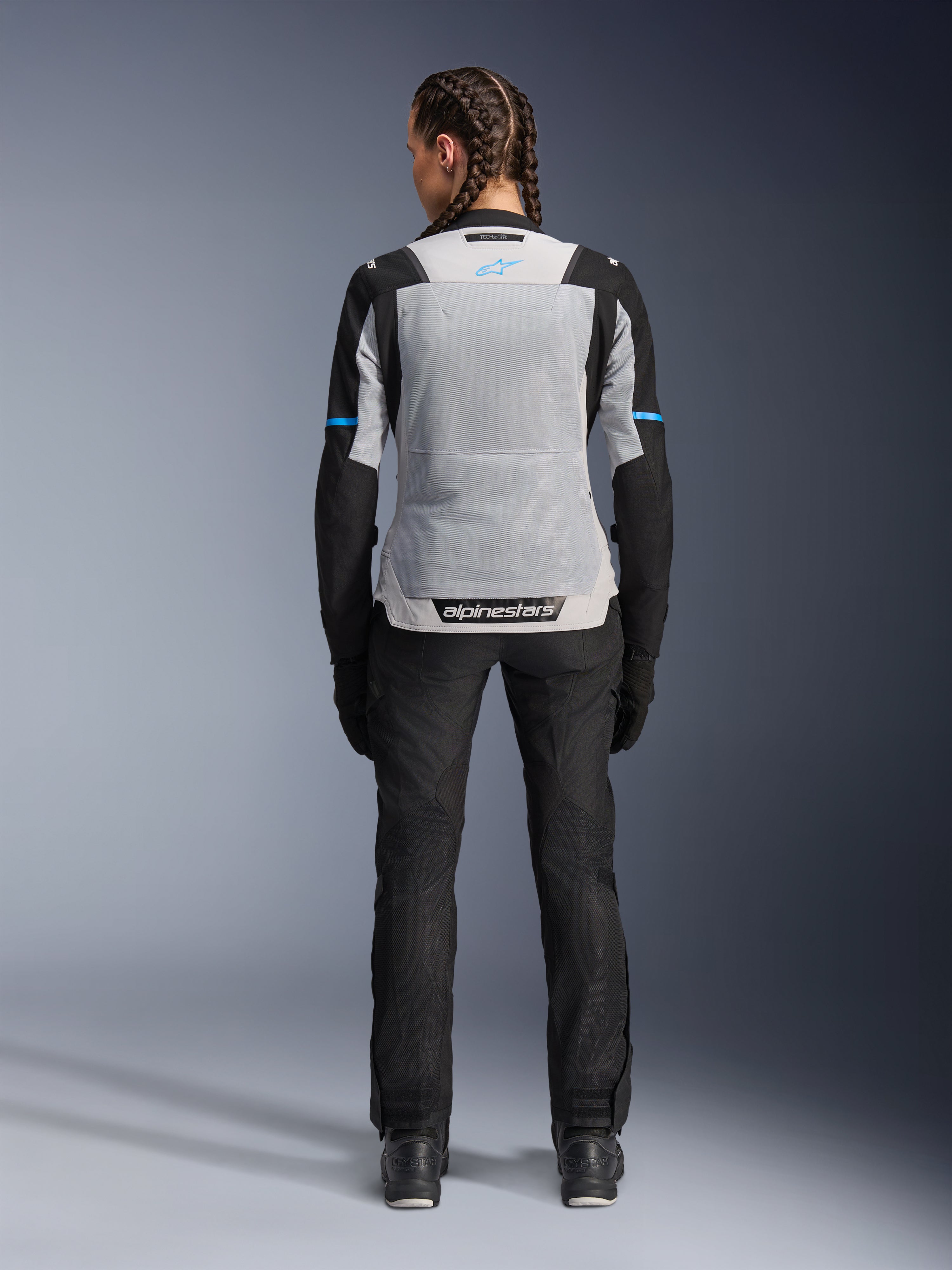 Chaqueta Alpinestars Stella ST-2 Air, Ice Gray Black, vista trasera en modelo femenina, con un gran panel de malla gris claro en la espalda, mangas negras con detalles de franjas azules, logotipo de Alpinestars en la parte inferior de la espalda, combinada con pantalones técnicos de conducción y botas negras, equipamiento de touring de verano para moto