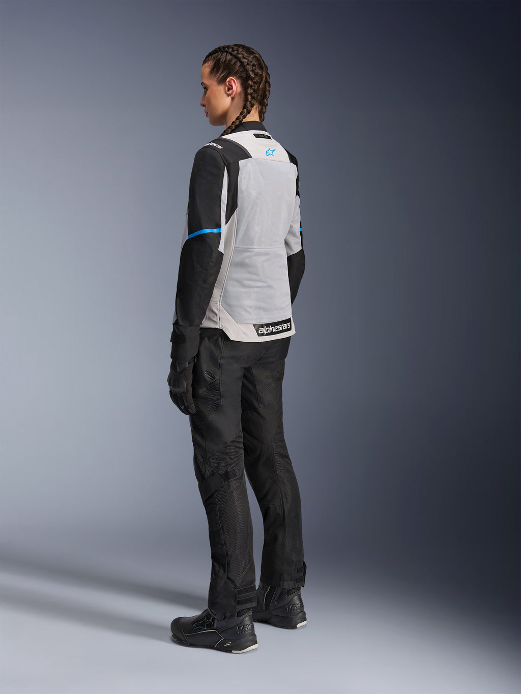 Mujer piloto con la chaqueta Alpinestars Stella ST-2 Air en Ice Gray y negro, vista trasera de tres cuartos que resalta el gran panel de malla gris claro en la espalda, mangas negras con una franja azul y el logotipo de Alpinestars en azul, combinada con pantalones, guantes y botas de moto negros para un conjunto completo de adventure touring