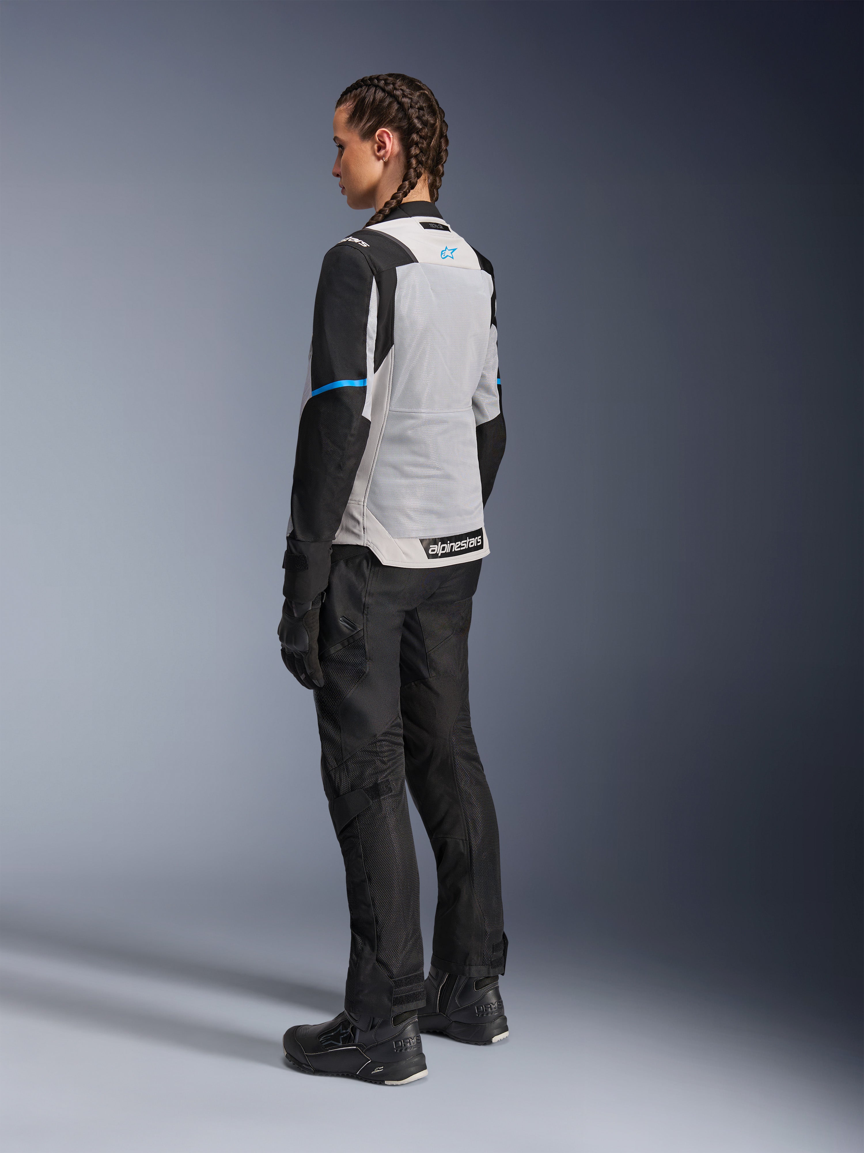 Mujer piloto con la chaqueta Alpinestars Stella ST-2 Air en Ice Gray y negro, vista trasera de tres cuartos que resalta el gran panel de malla gris claro en la espalda, mangas negras con una franja azul y el logotipo de Alpinestars en azul, combinada con pantalones, guantes y botas de moto negros para un conjunto completo de adventure touring
