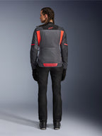 Vista trasera de una modelo con la chaqueta Alpinestars Stella St-2 Air para mujer en Deep Gray y Dark Gray con detalles en Fluo Coral, que presenta grandes paneles de ventilación de malla en la espalda y detalles de logotipos reflectantes, combinada con pantalones de moto y guantes negros.