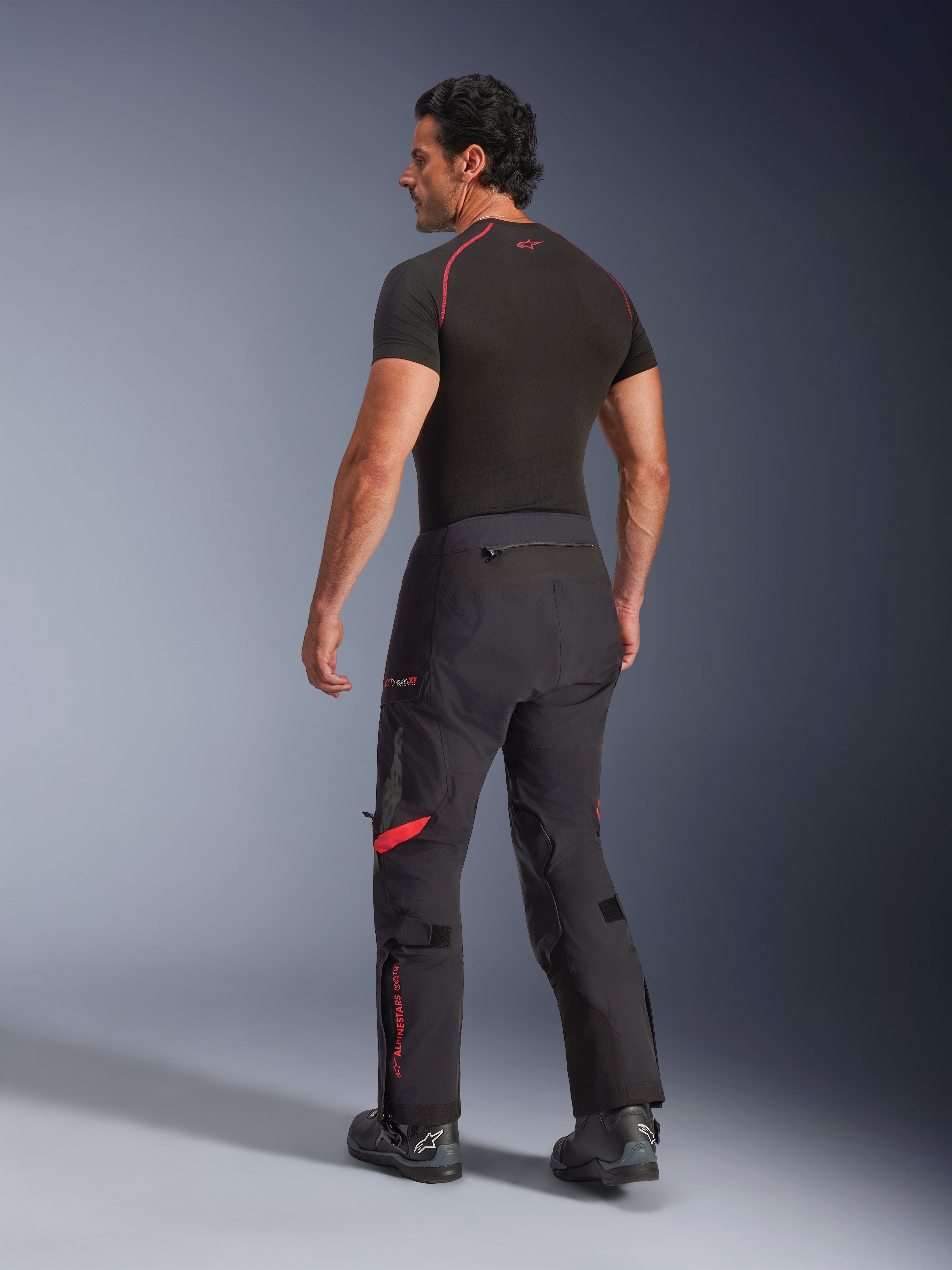 Pantalones laminados Alpinestars Halo Pro Drystar® XF, color Black Dark Shadow con detalles rojos, vista trasera en modelo masculino, combinados con una capa base negra y botas de moto, equipo técnico de aventura para conducción todoterreno