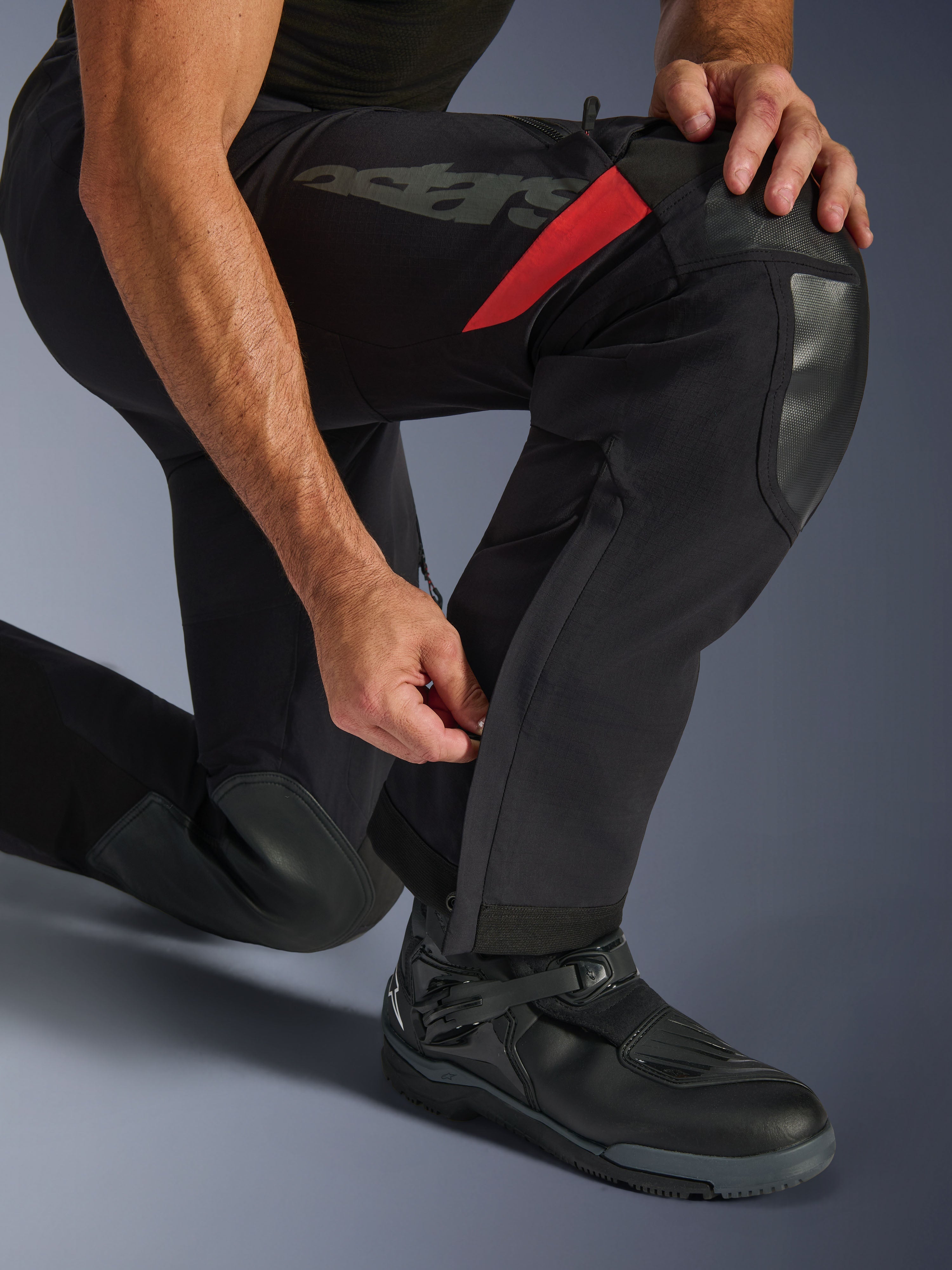 Pantalones laminados Alpinestars Halo Pro Drystar® XF, Black Dark Shadow, primer plano de la parte inferior de la pierna con cremallera ajustable en la pantorrilla, con refuerzos duraderos en las rodillas, detalles en rojo y marca 'astars', usados con botas de moto negras.
