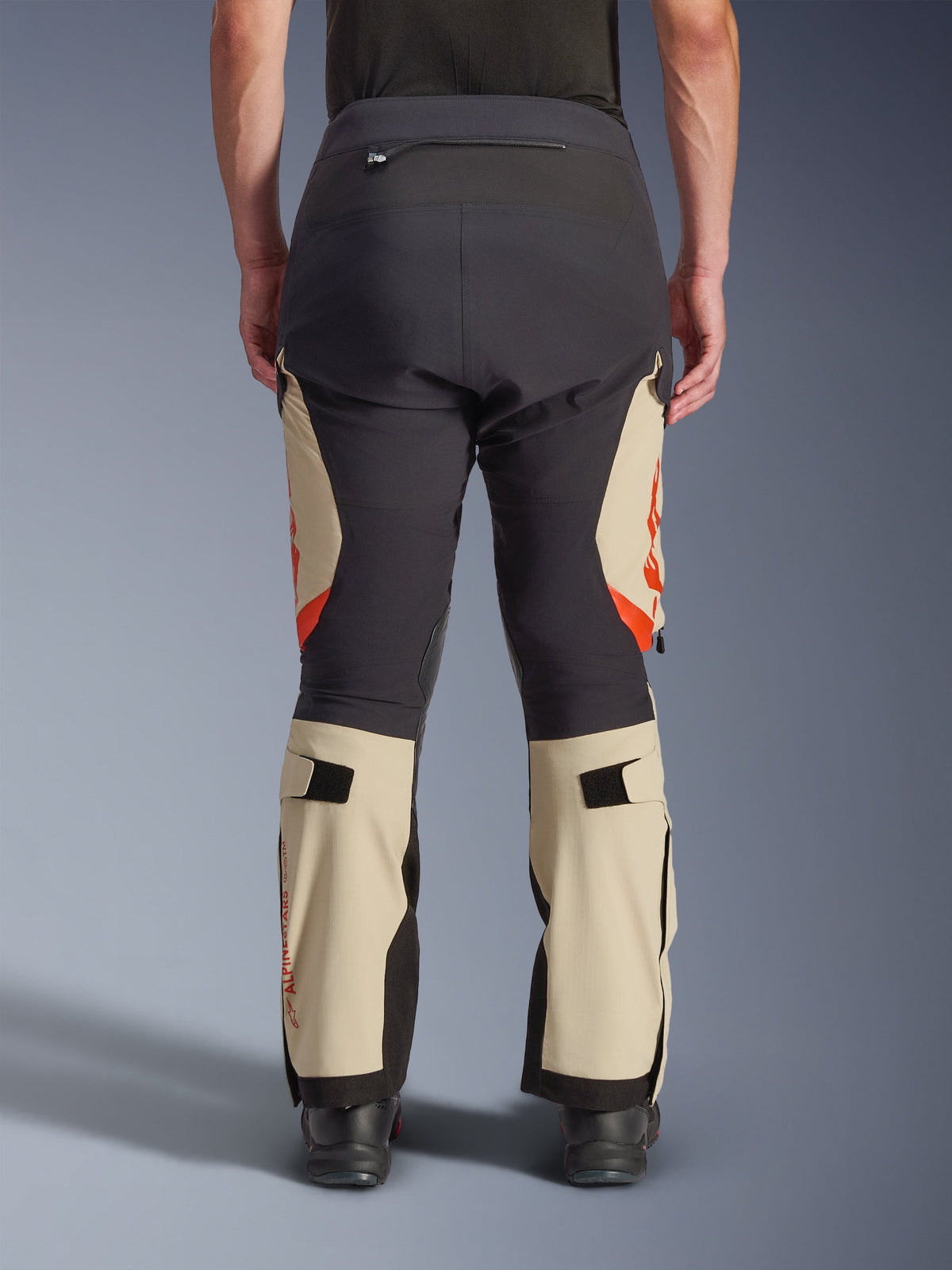 Vista trasera de los pantalones laminados Alpinestars Halo Pro Drystar® XF, Mastic Black con paneles beige y detalles naranjas, pantalones de motociclismo de aventura con construcción textil técnica, usados con botas de motociclismo negras