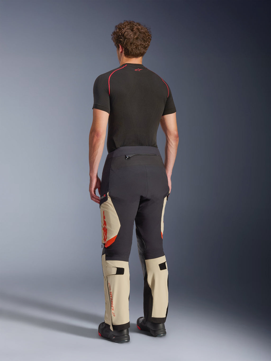 Pantalones laminados Alpinestars Halo Pro Drystar® XF, pantalones de moto de aventura, negro Mastic y tostado con detalles en naranja, vista trasera que muestra la construcción de la capa laminada de 3 capas, combinados con una capa base negra y botas de aventura.
