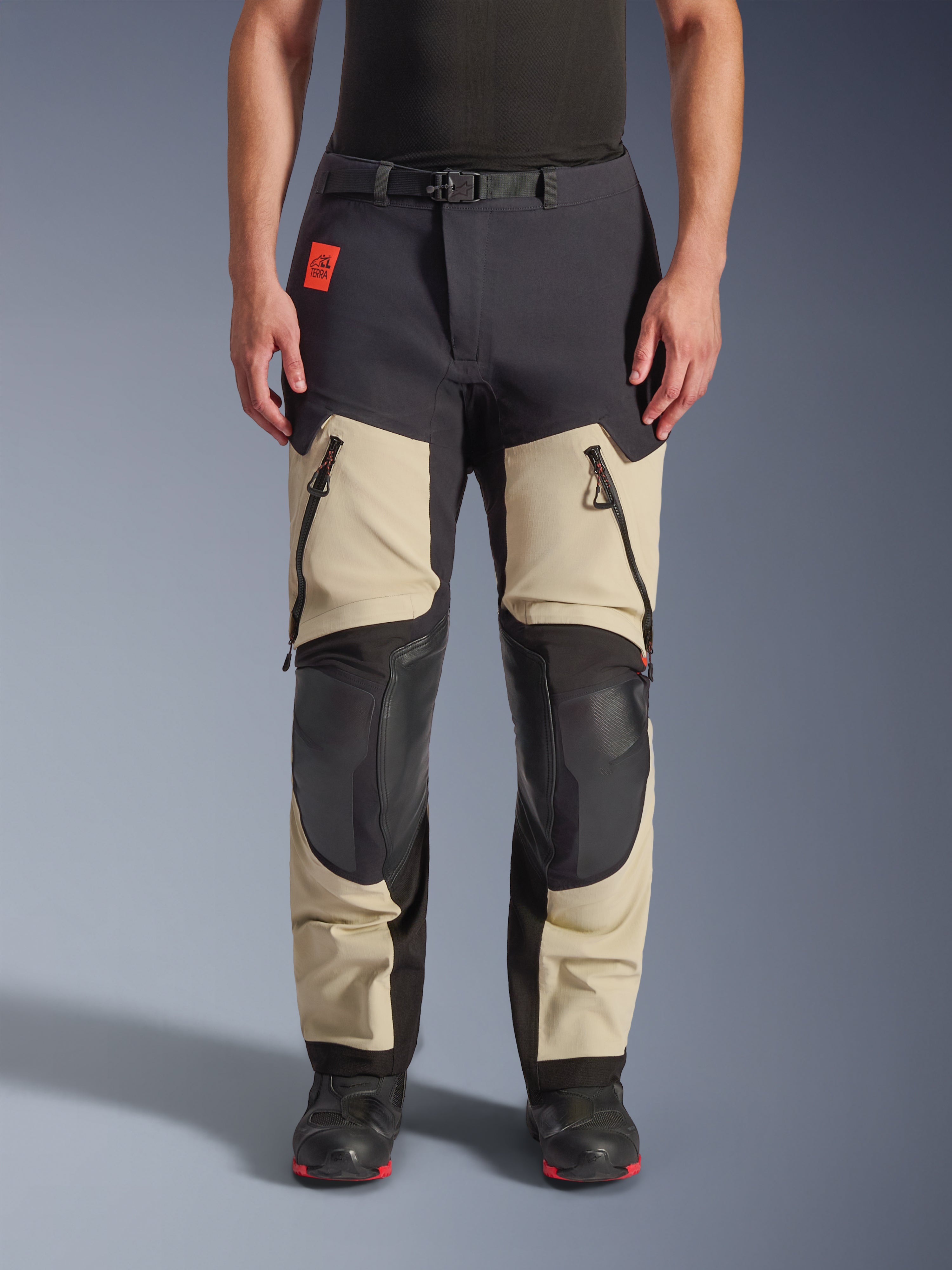 Pantalones laminados Alpinestars Halo Pro Drystar® XF, pantalones de moto de aventura, Mastic Black, vista frontal en un piloto, con grandes ventilaciones con cremallera en los muslos, paneles reforzados negros en las rodillas y confección textil en color tan, combinados con botas de moto negras y rojas.