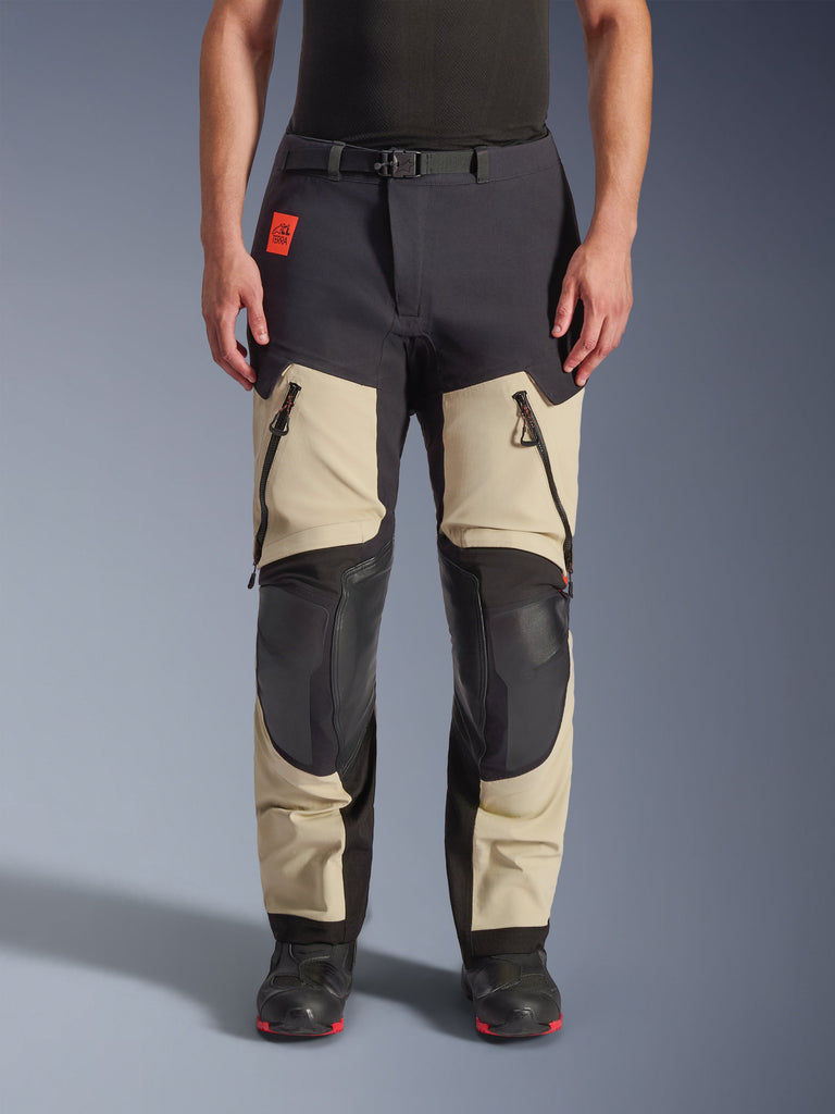 Pantalones laminados Alpinestars Halo Pro Drystar® XF, pantalones de moto de aventura, Mastic Black, vista frontal en un piloto, con grandes ventilaciones con cremallera en los muslos, paneles reforzados negros en las rodillas y confección textil en color tan, combinados con botas de moto negras y rojas.