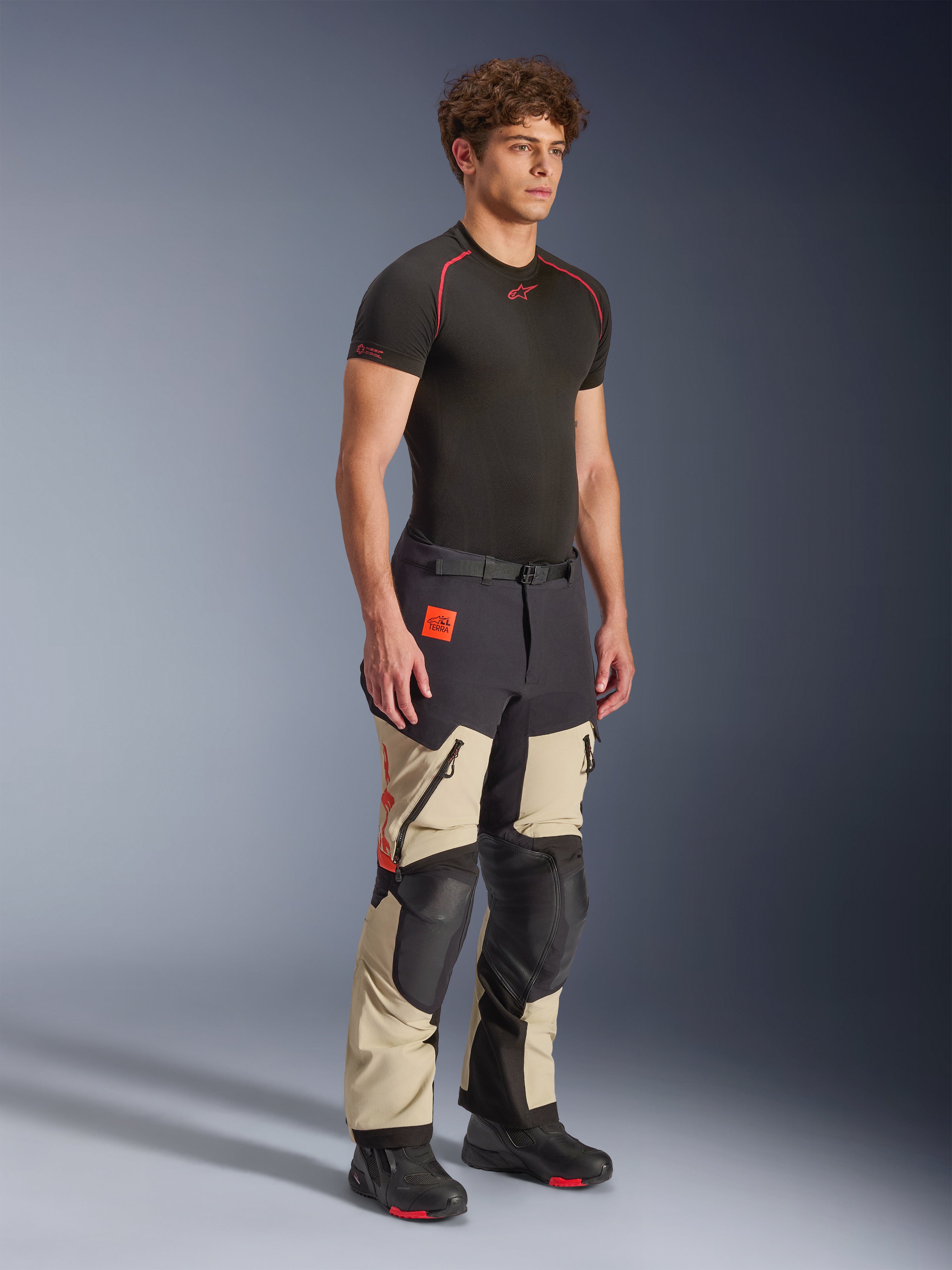 Pantalones laminados Alpinestars Halo Pro Drystar® XF en Mastic Black, pantalones de moto de aventura usados por un modelo, con refuerzos negros en las rodillas y el asiento, logotipos en naranja y múltiples cremalleras funcionales, combinados con una capa base negra y botas de moto.