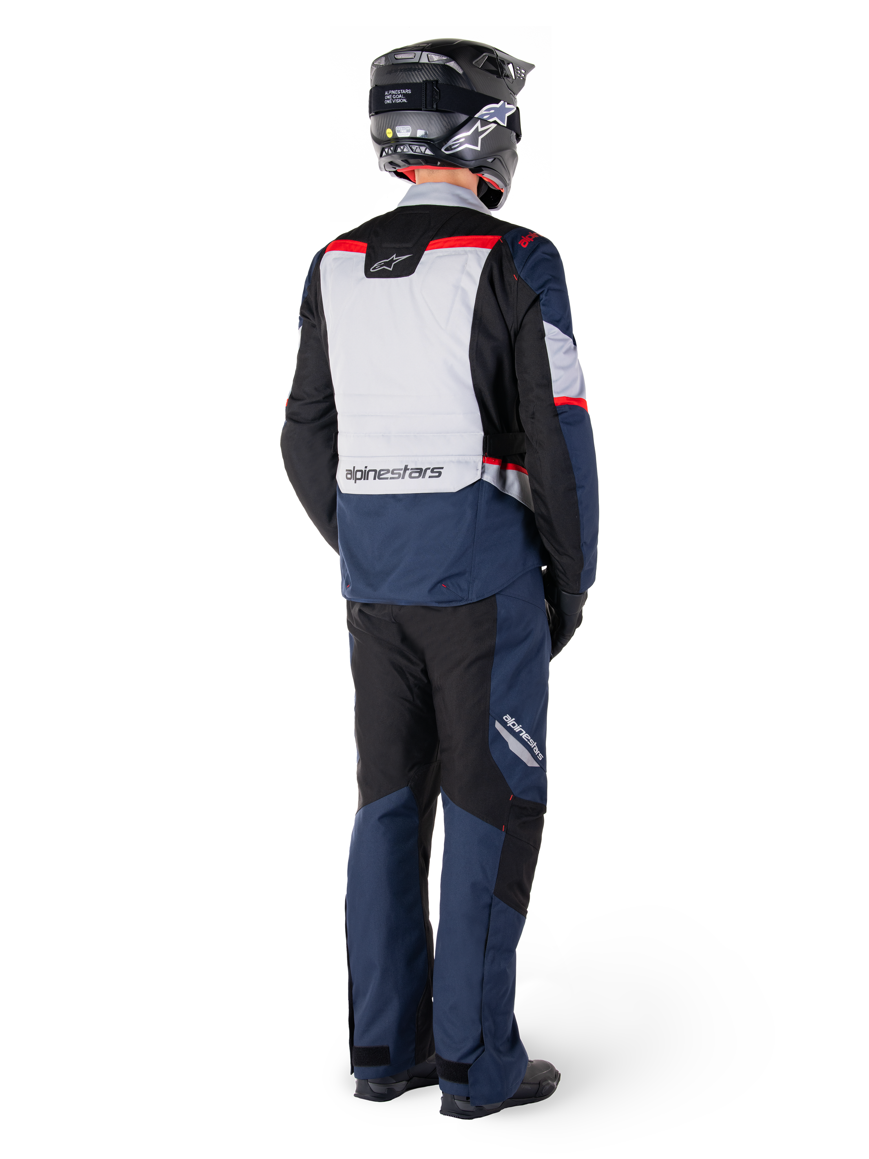 Vista trasera de un motociclista con pantalones Alpinestars ST-1 Waterproof y chaqueta a juego en azul marino, negro y rojo brillante, equipamiento de turismo de aventura con paneles textiles y detalles reflectantes, combinado con casco y botas negras.