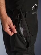 Primer plano de los pantalones Alpinestars Andes V4 Drystar® en color negro, que muestra una mano abriendo la ventilación directa en el muslo con forro de malla, resaltando la construcción textil duradera y el logotipo de Alpinestars.