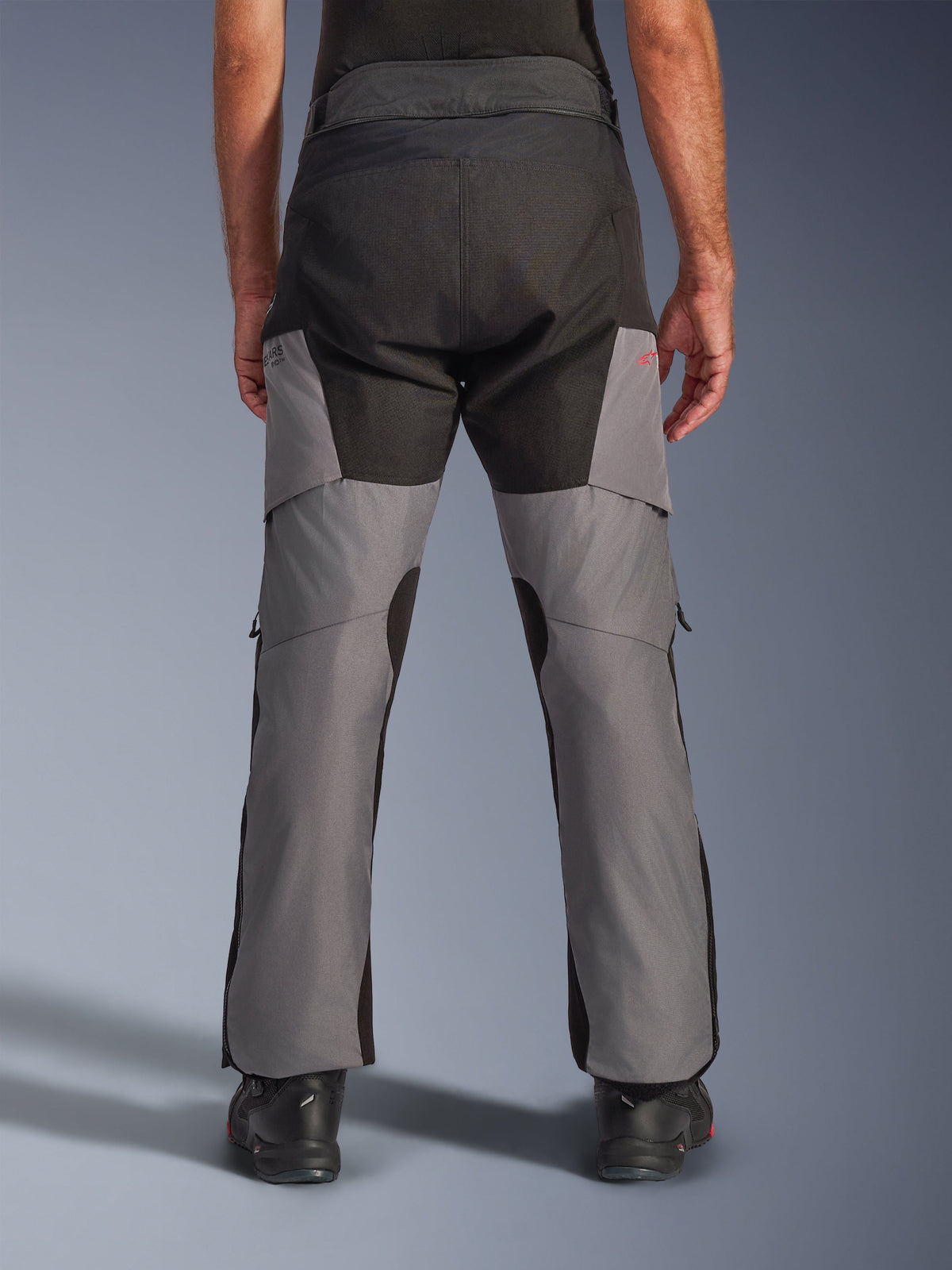 Pantalones Alpinestars Andes V4 Drystar®, pantalones de moto de aventura, negro y gris oscuro, vista trasera que muestra el asiento negro reforzado y las piernas grises, con un pequeño logo rojo, combinados con botas de motociclismo negras, construcción textil duradera para touring en cualquier clima.