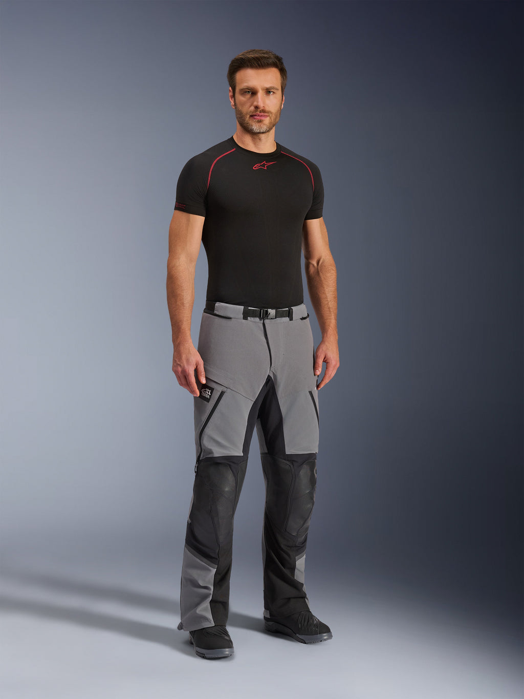 Pantalones Alpinestars Andes Pro Drystar® XF, pantalones de aventura para motociclismo, gris oscuro y negro, vestidos por un modelo con una capa base técnica negra, con construcción de múltiples paneles, cremalleras impermeables y zonas reforzadas para conducción de aventura.