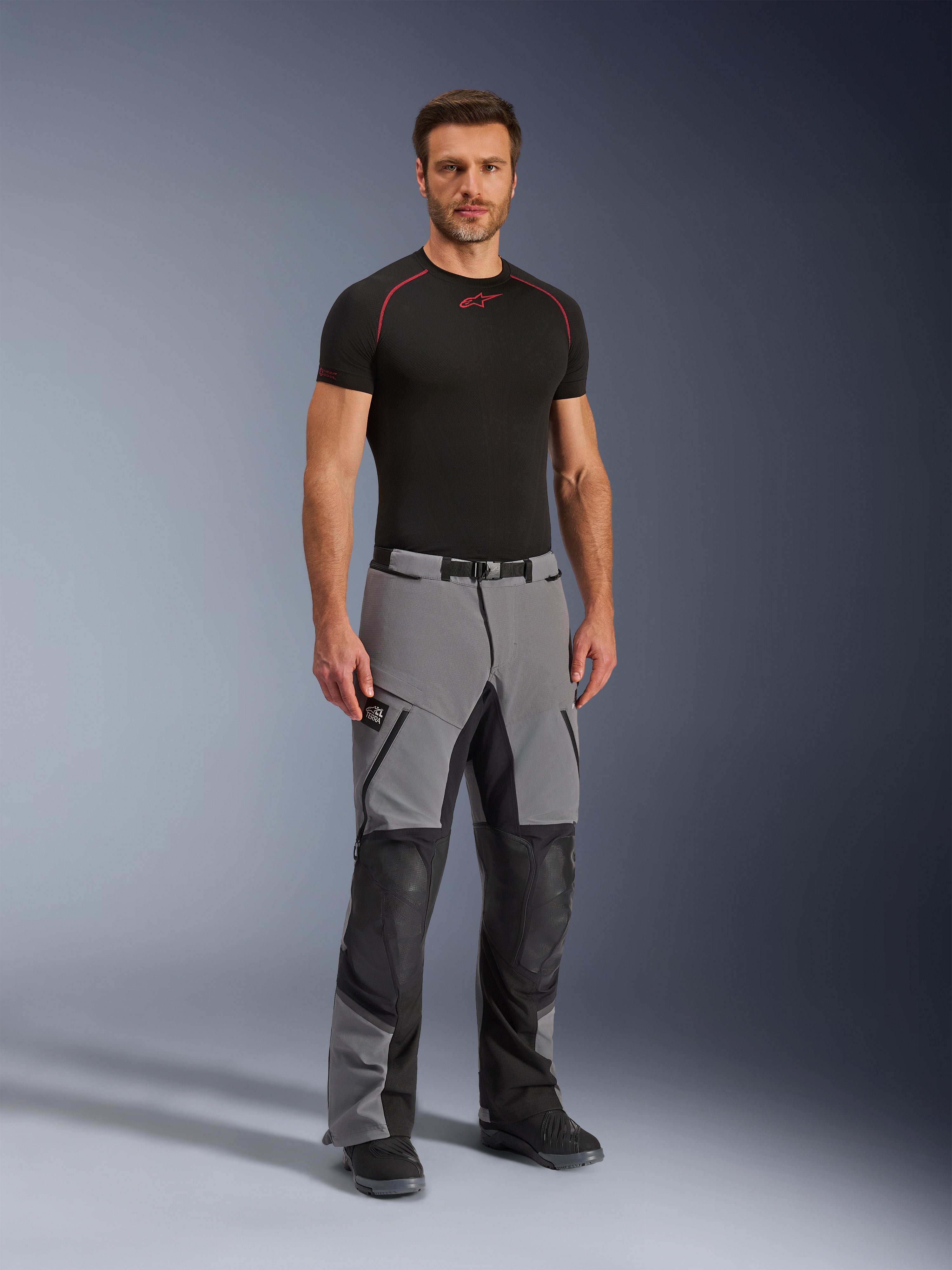 Pantalones Alpinestars Andes Pro Drystar® XF, pantalones de aventura para motociclismo, gris oscuro y negro, vestidos por un modelo con una capa base técnica negra, con construcción de múltiples paneles, cremalleras impermeables y zonas reforzadas para conducción de aventura.