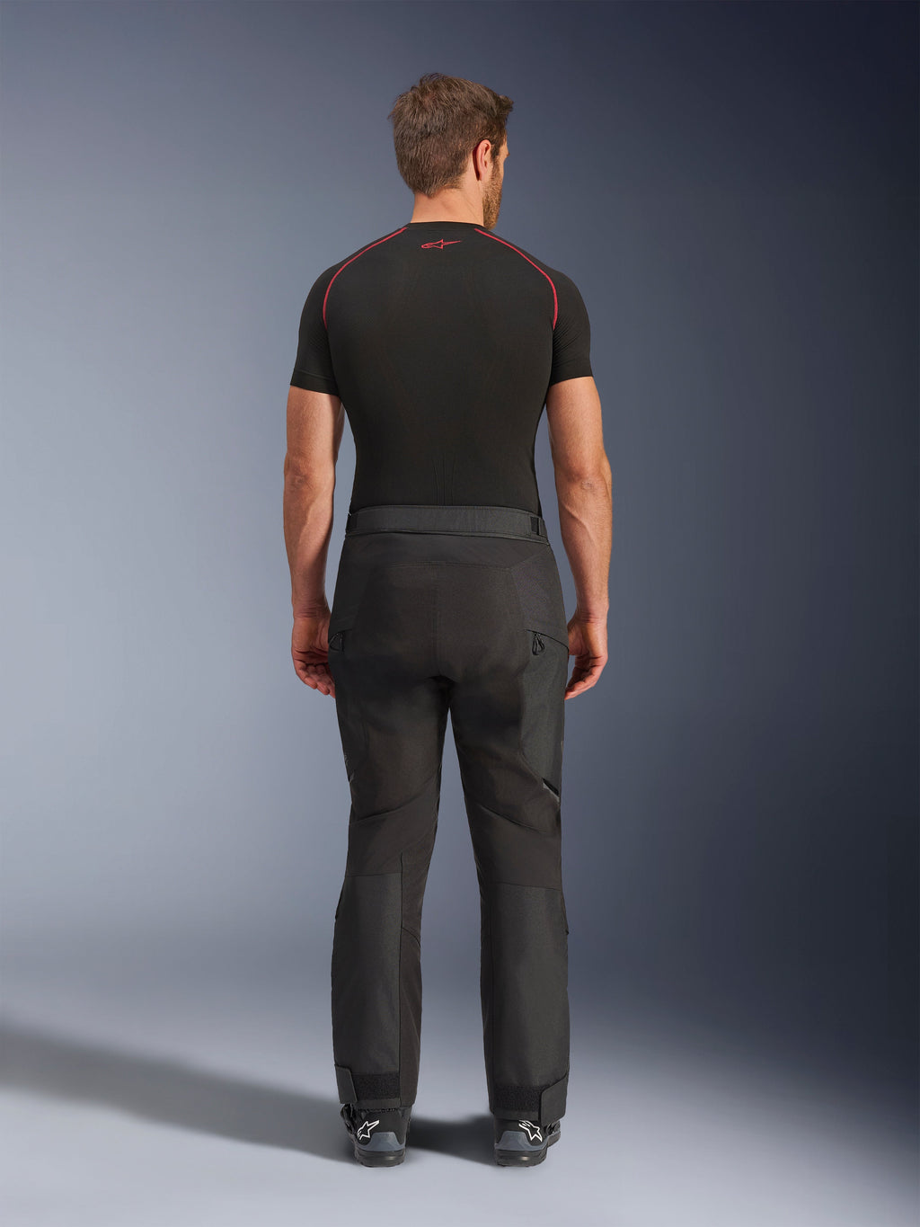 Motociclista con pantalones Alpinestars Cusco Drystar®, pantalones de motociclismo adventure negros, vista trasera, combinados con una camiseta de compresión negra de manga corta y botas de motociclismo negras, con un diseño textil duradero y paneles articulados para touring