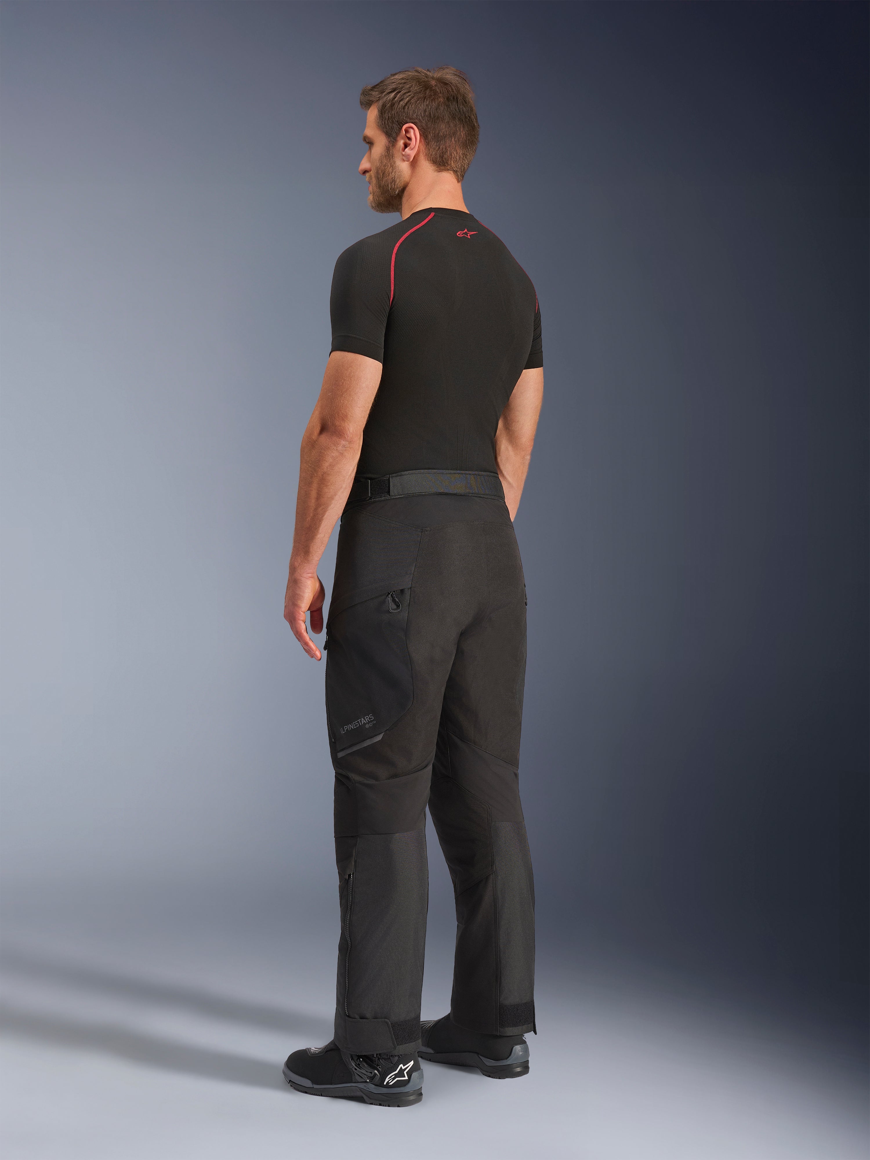 Pantalones Alpinestars Cusco Drystar®, pantalones de aventura para moto, negros, usados por un modelo en una vista lateral y trasera, combinados con una capa base negra y botas de moto negras, construcción textil técnica con bolsillos con cremallera y rodillas articuladas