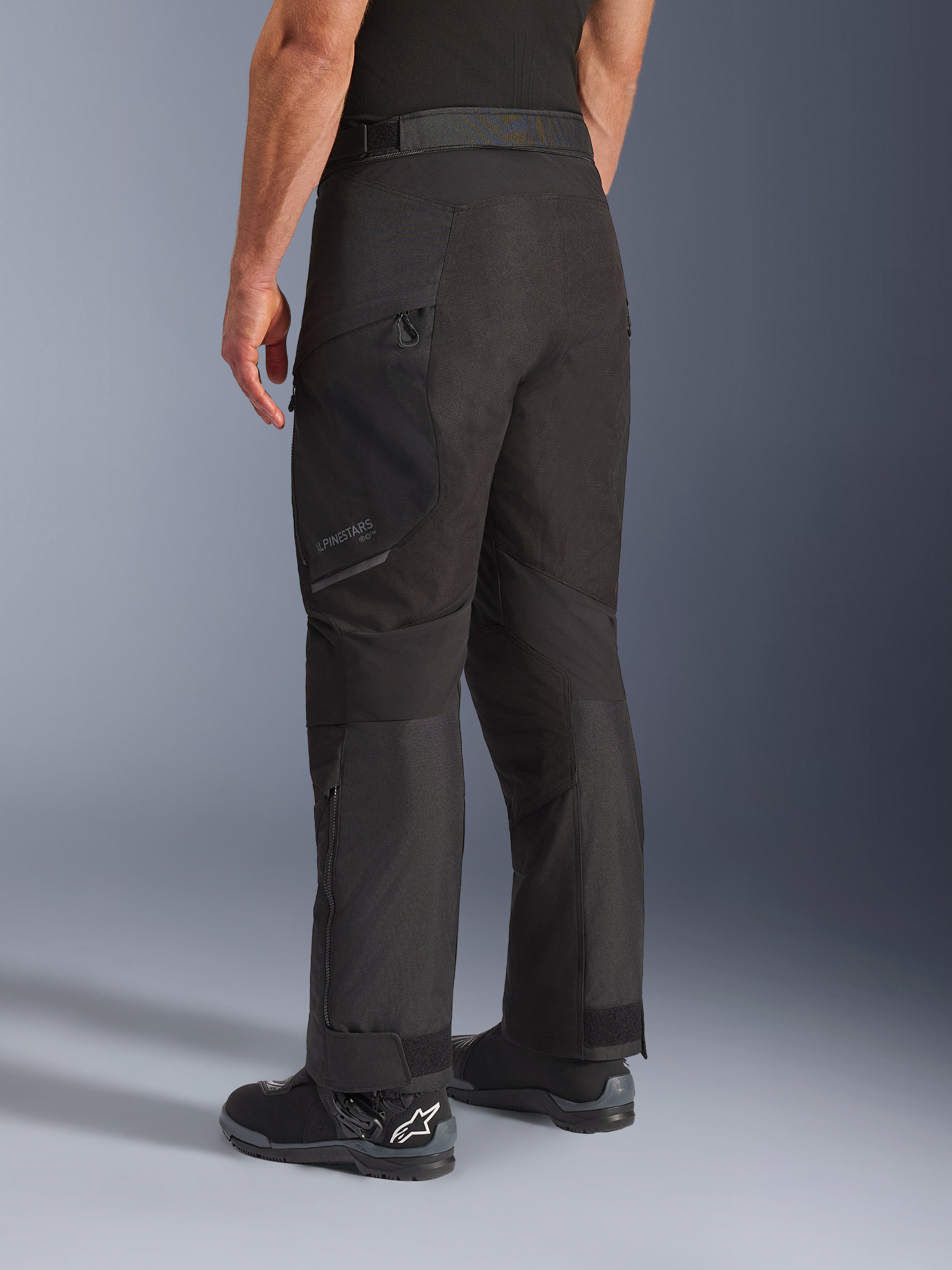 Pantalones Alpinestars Cusco Drystar®, pantalones de moto, negro, vista trasera de tres cuartos que muestra el bolsillo con cremallera en el muslo y paneles articulados, combinados con botas de moto negras, diseño duradero de adventure touring con logotipos sutiles
