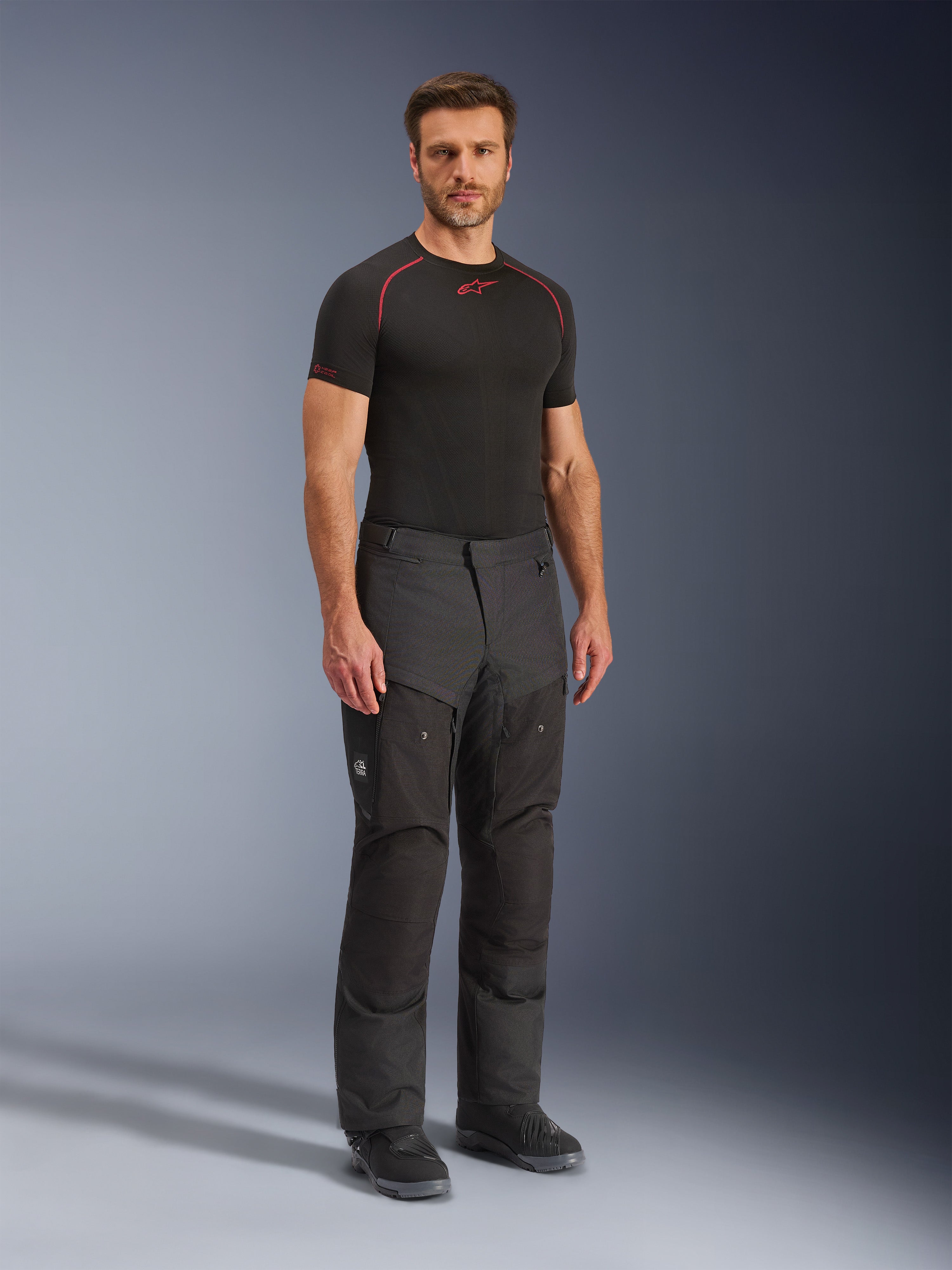 Pantalones Alpinestars Cusco Drystar®, pantalones de moto textiles negros, usados por un modelo con una capa base negra y botas, con tejido ripstop, bolsillos cargo grandes con la marca 'Terra' y diseño ergonómico para conducción de aventura.